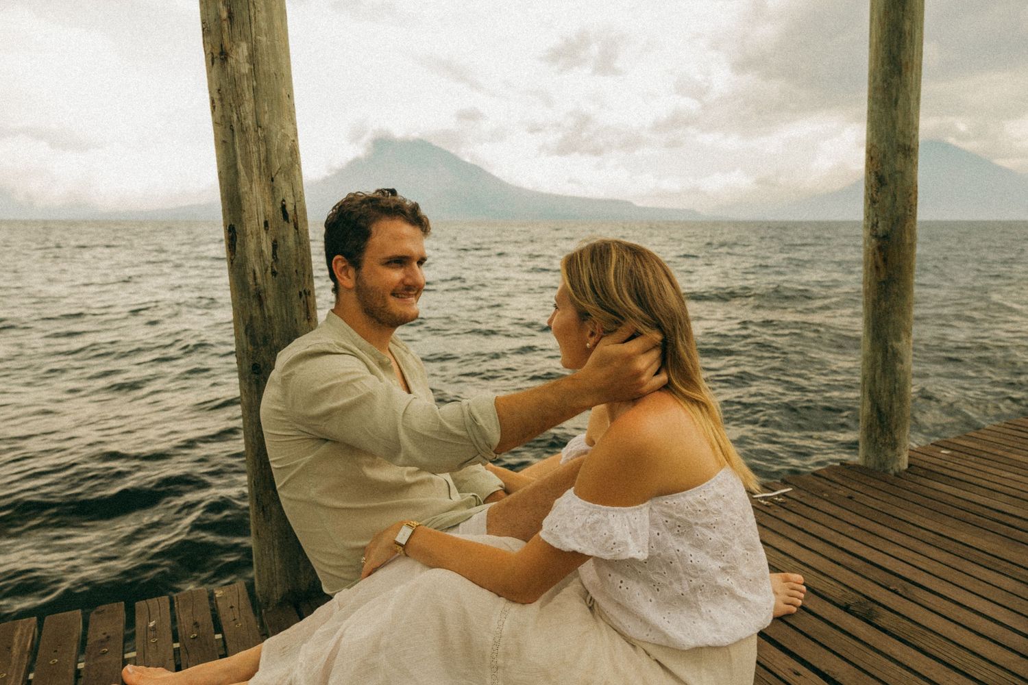 Couple sitting on a wooden dock by a lake with a volcano in the background on a cloudy day.