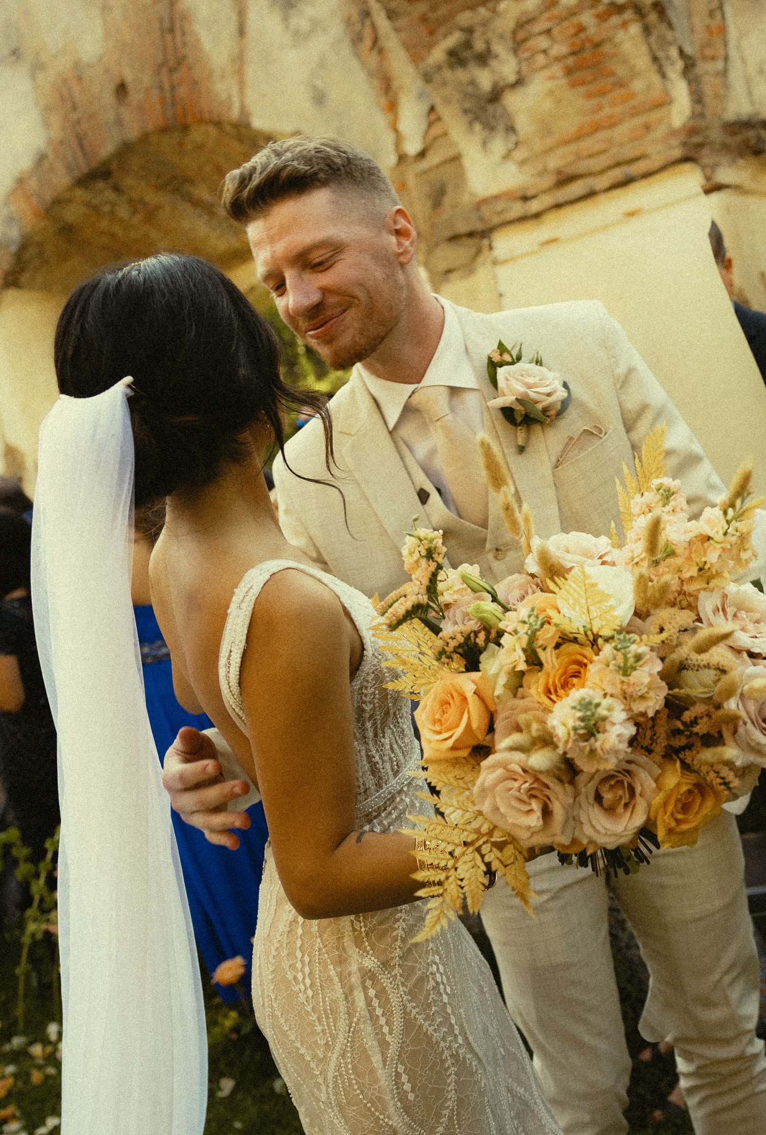 Bride in beaded gown holding peach floral bouquet with groom in tan suit smiling at outdoor wedding ceremony.