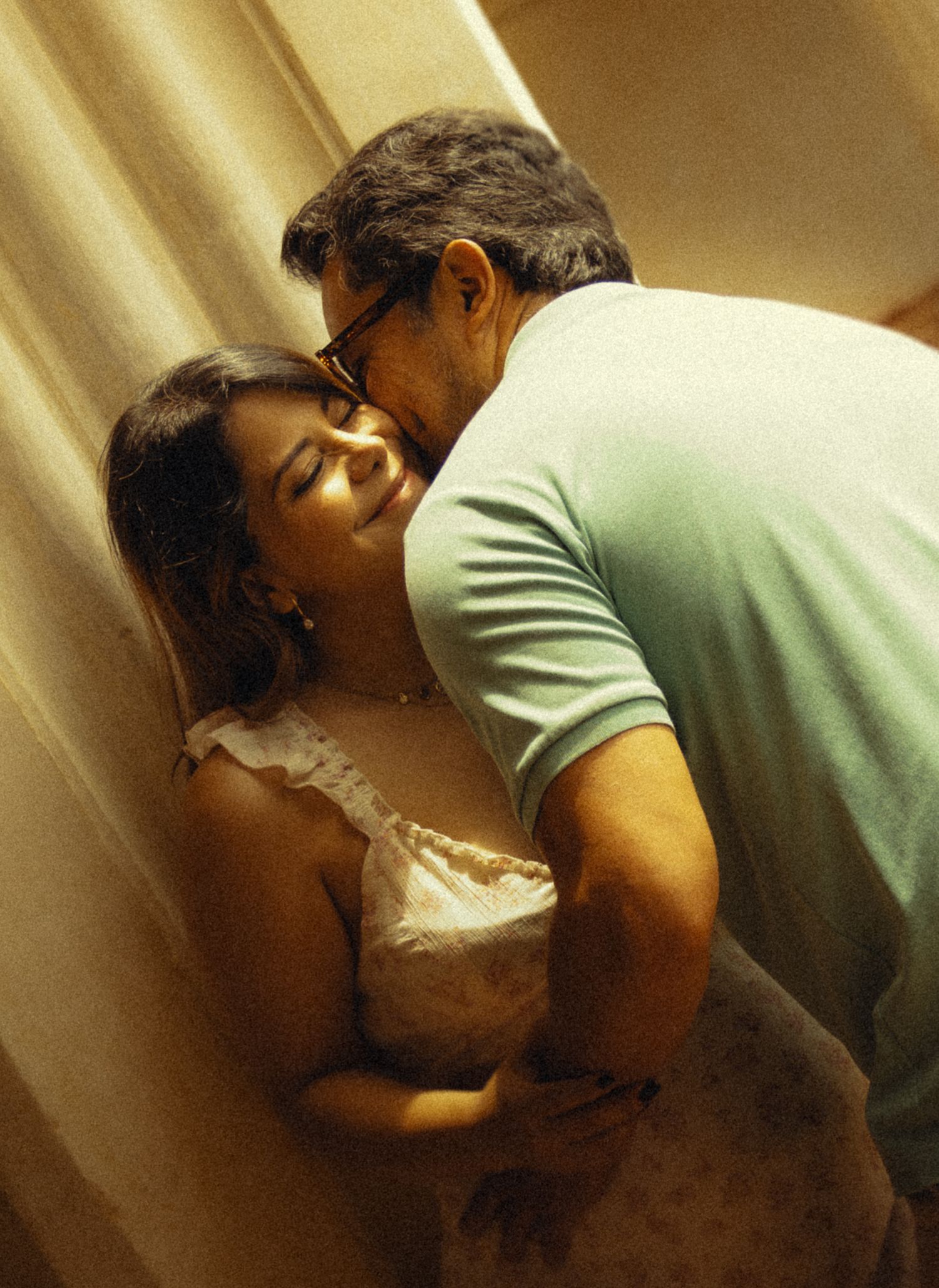 A couple sharing an intimate embrace near a curtain, man kissing woman's cheek softly.