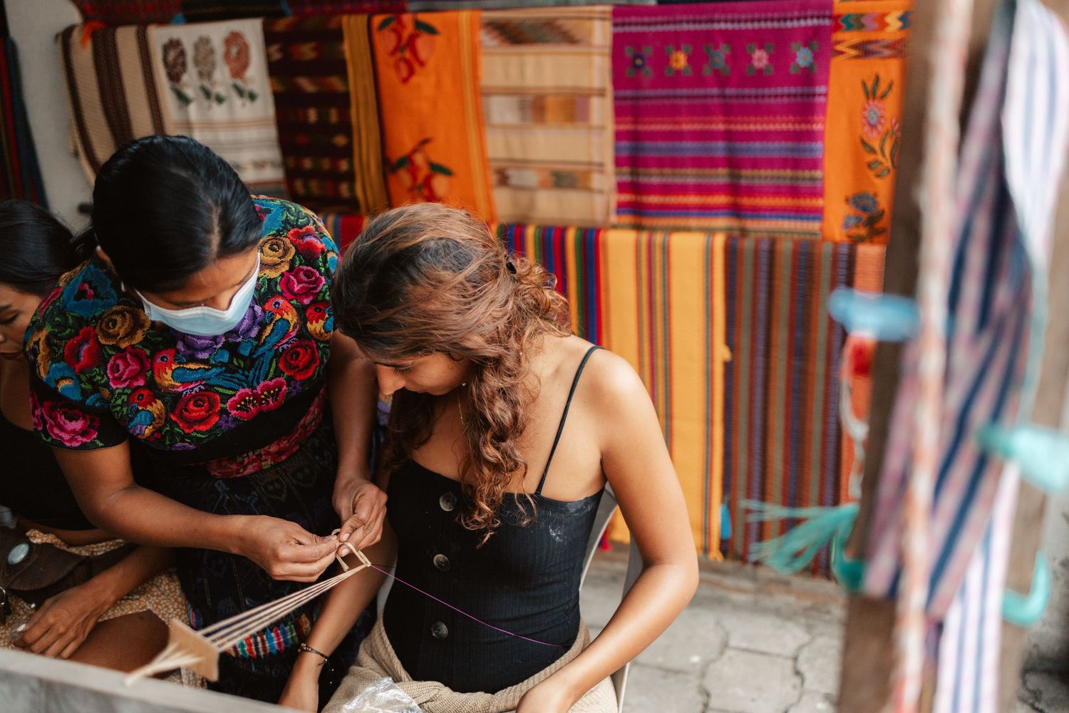 Two women examining traditional textile weaving at a colorful fabric market stall.