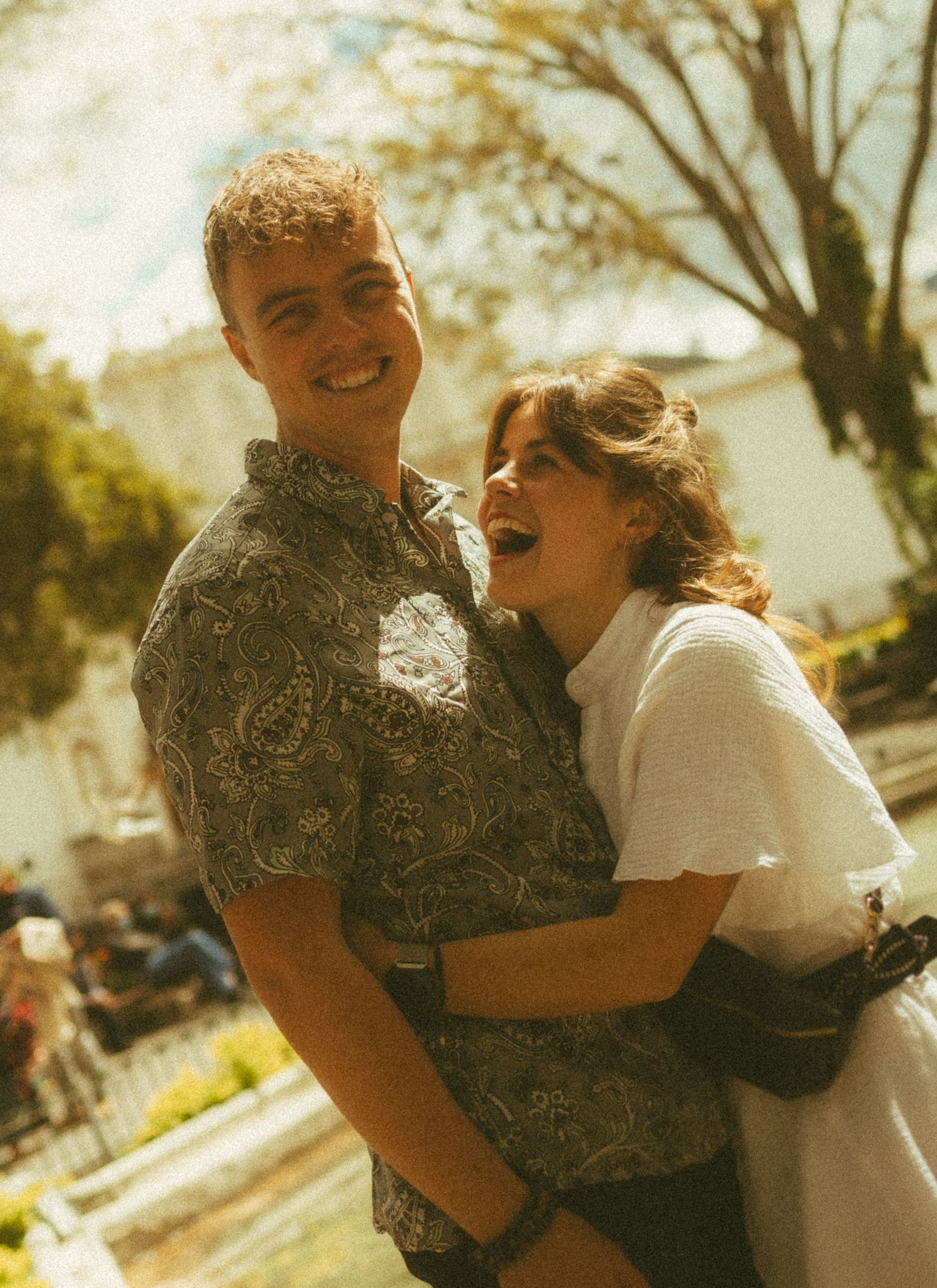 A smiling couple embracing outdoors on a sunny day, man in patterned shirt, woman in white top, trees in background.