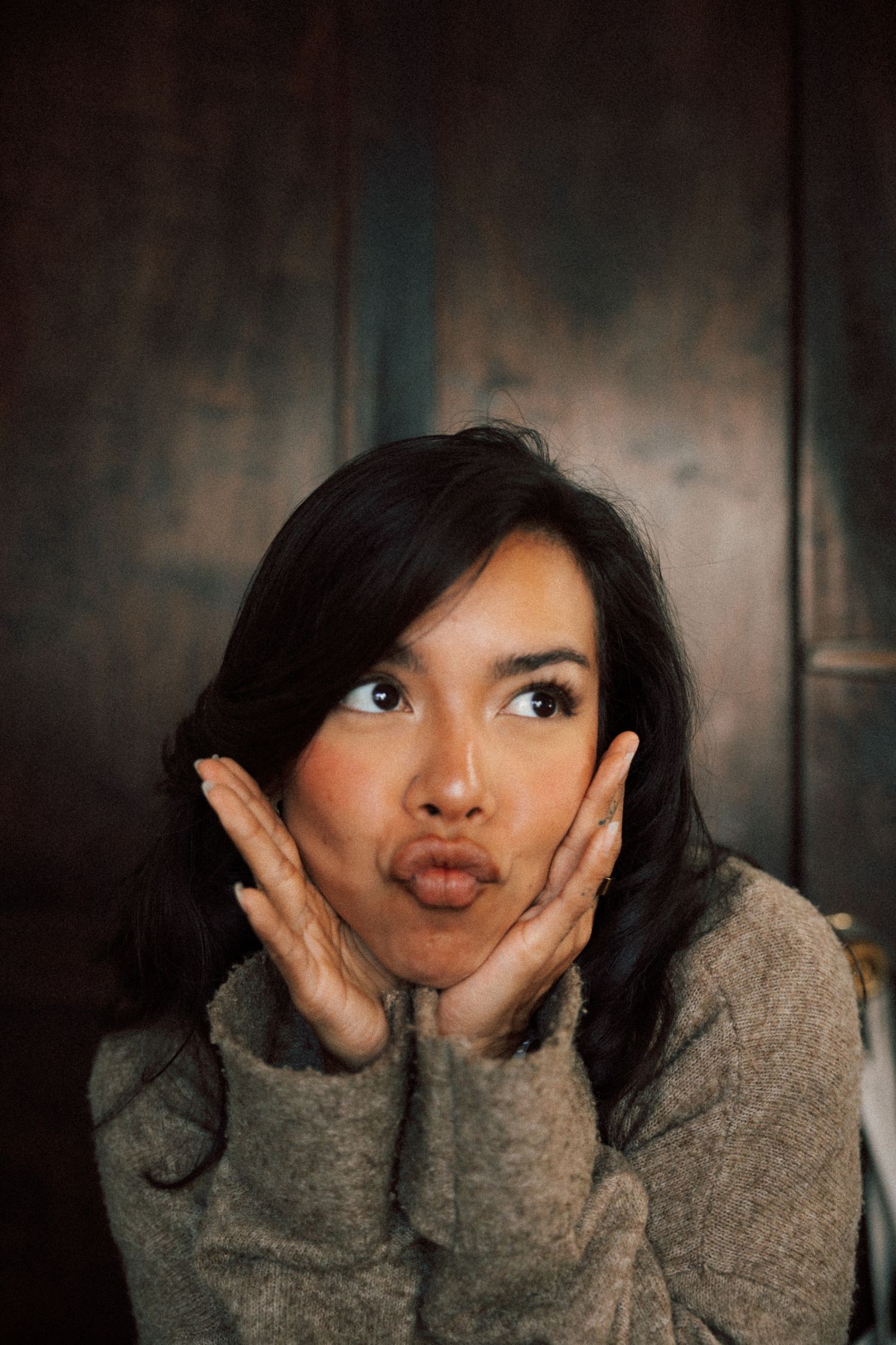 Young woman in gray sweater making a playful pouty face with hands on cheeks against dark wood background.