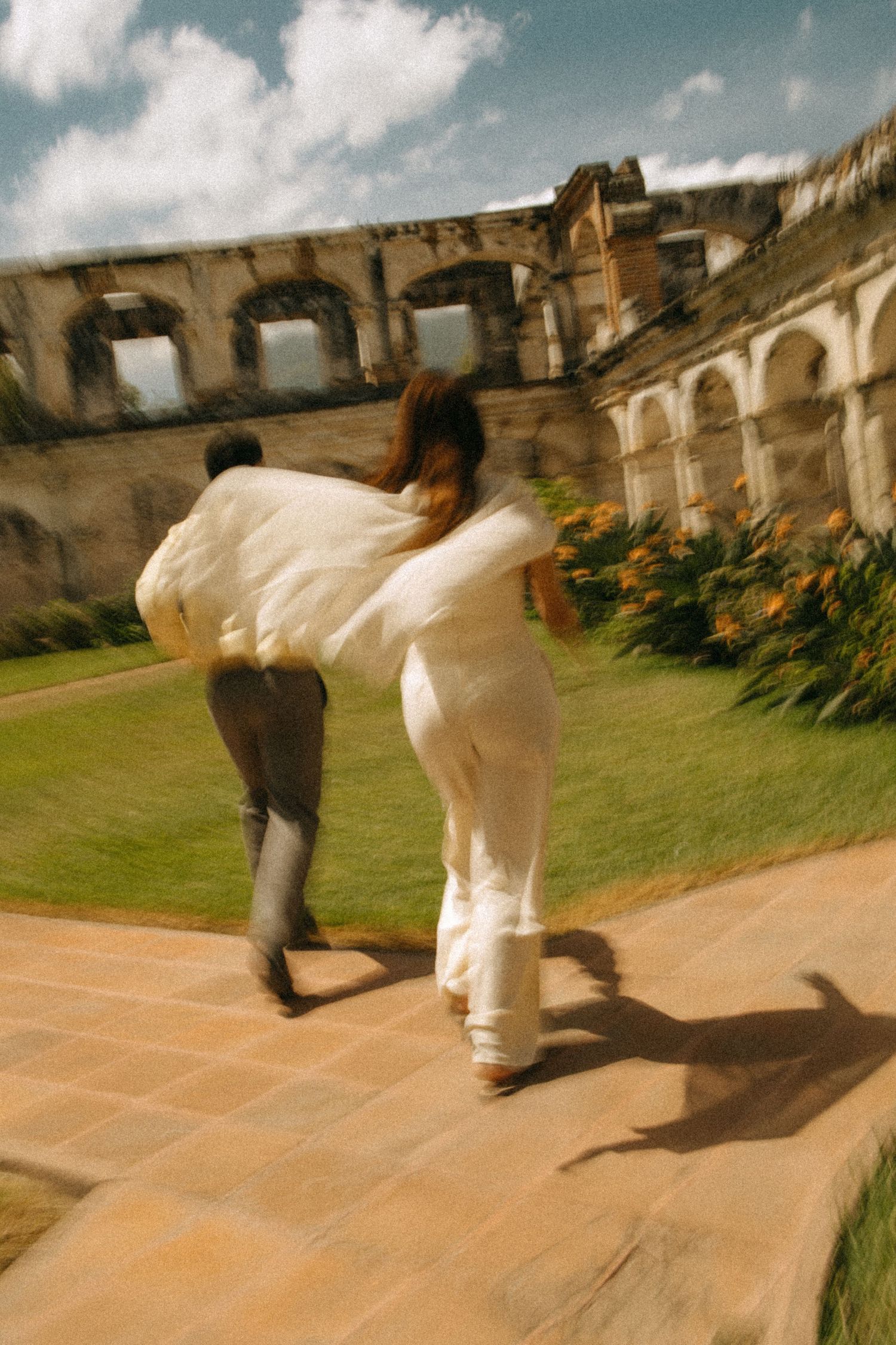 Couple in white walks hand in hand across a sunlit brick path past a lush green lawn and historic stone ruins with arched windows.