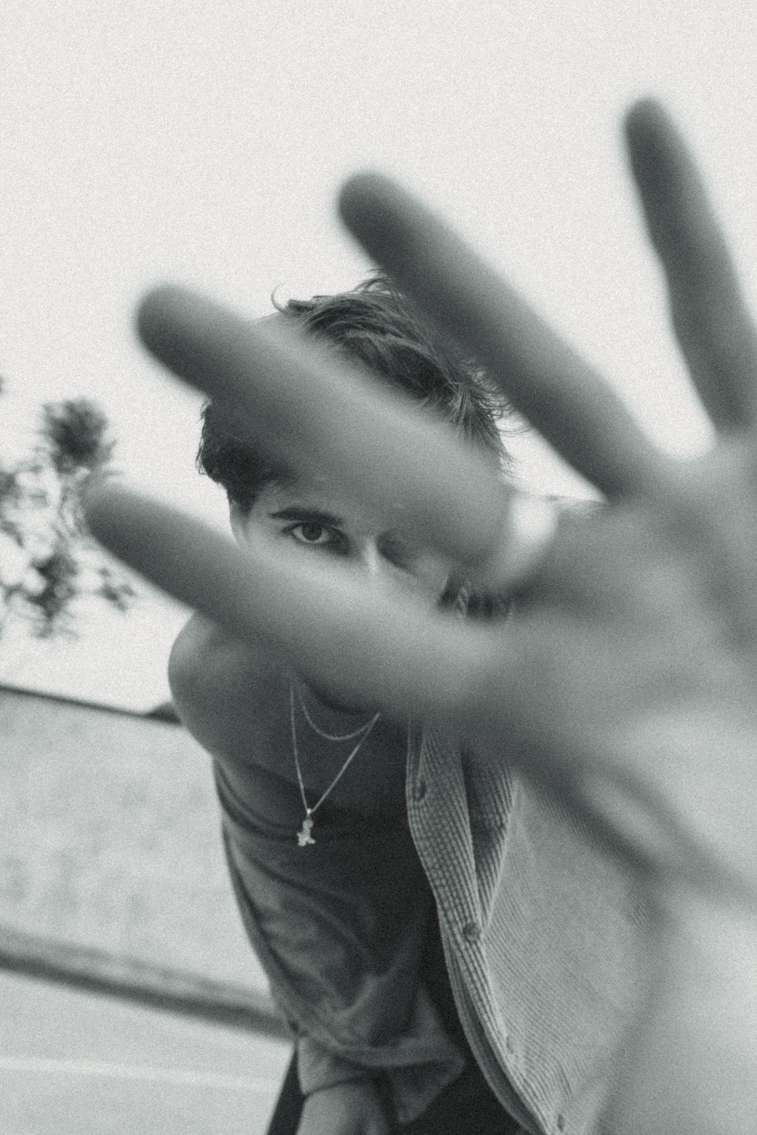 Black and white photo of a young man extending his hand toward the camera, wearing layered necklaces outdoors.