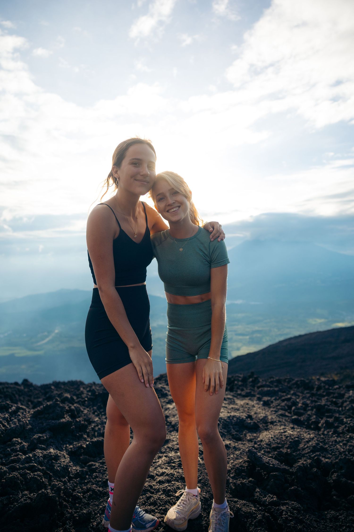 Two women in athletic wear posing together on a volcanic mountain summit with scenic sky views.