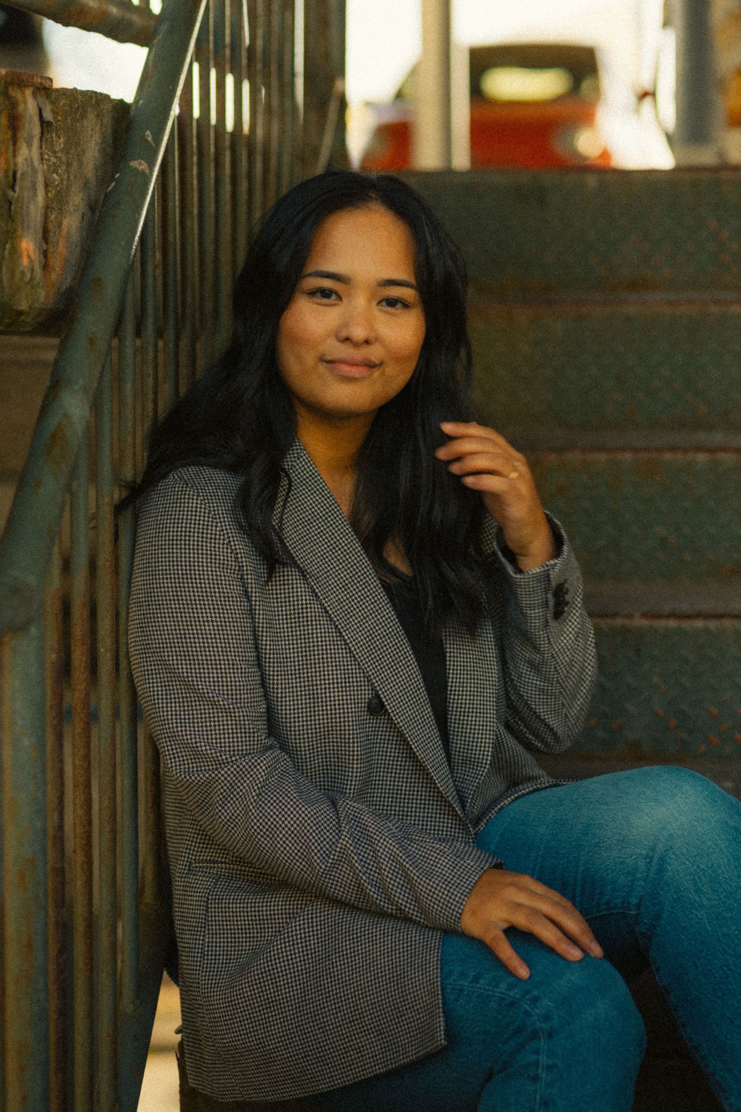 Young woman in gray blazer and jeans sitting on metal steps, smiling softly at camera.