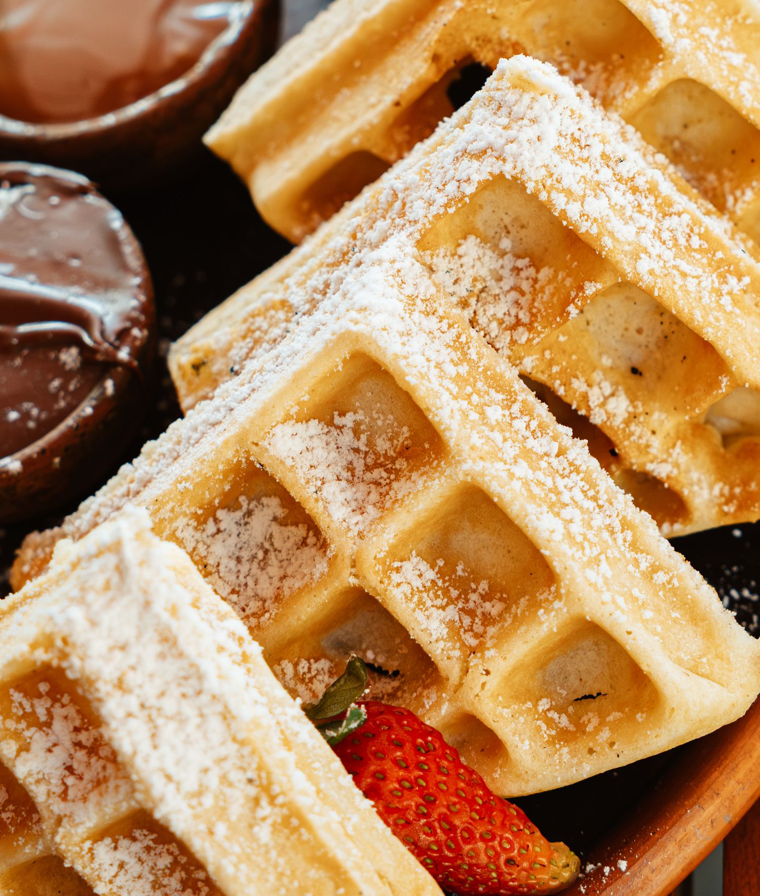 Golden Belgian waffles dusted with powdered sugar, served with fresh strawberries and chocolate dipping sauce on a wooden plate.