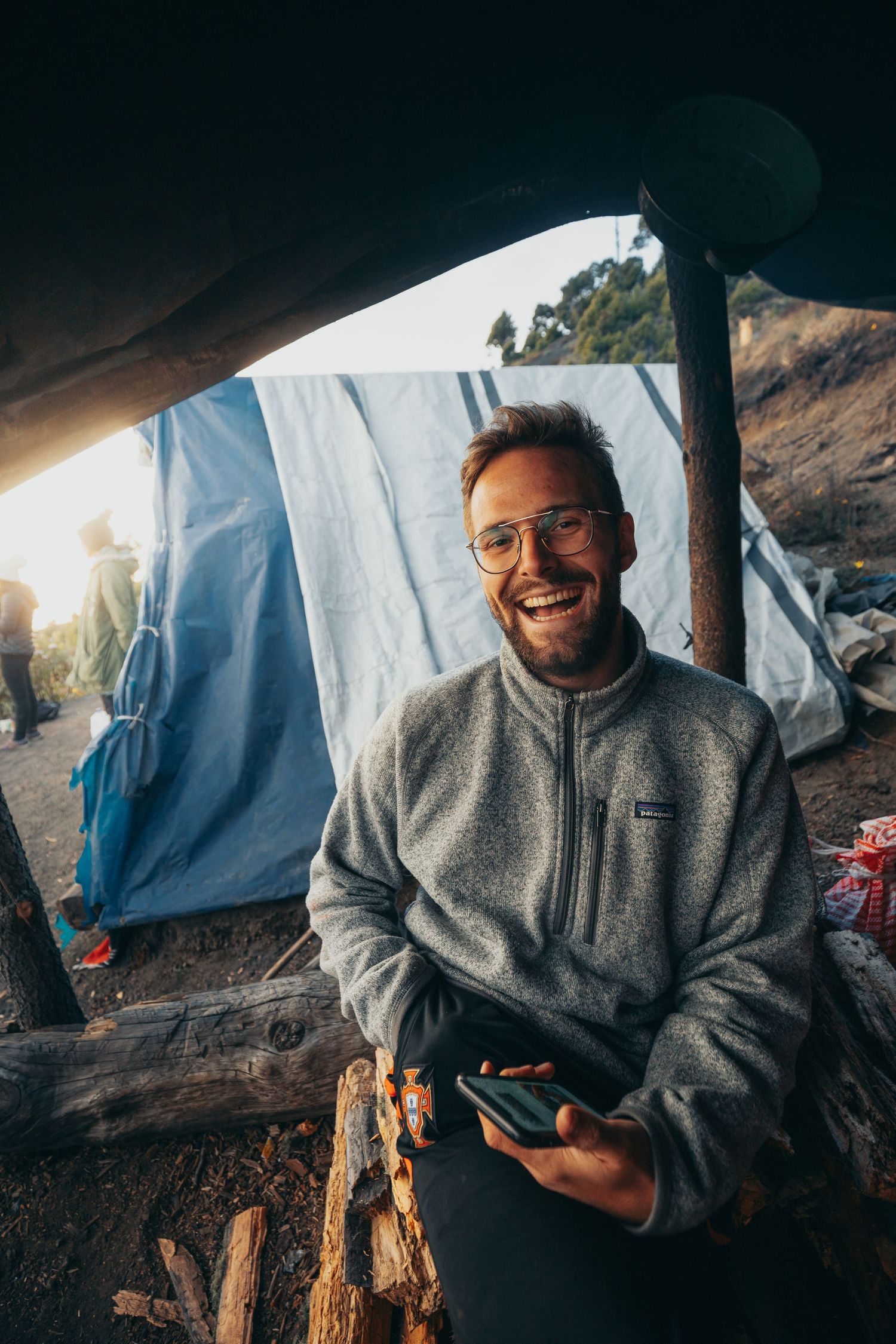 Happy man in gray Patagonia fleece holding phone while sitting near a tent at a mountain campsite.
