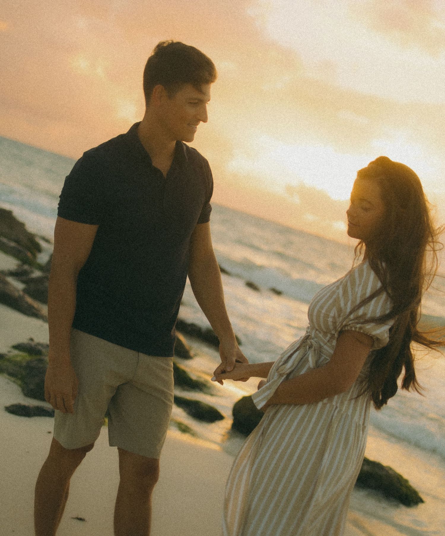 Couple holding hands on a sandy beach at sunset, man in polo shirt and shorts, woman in striped dress.