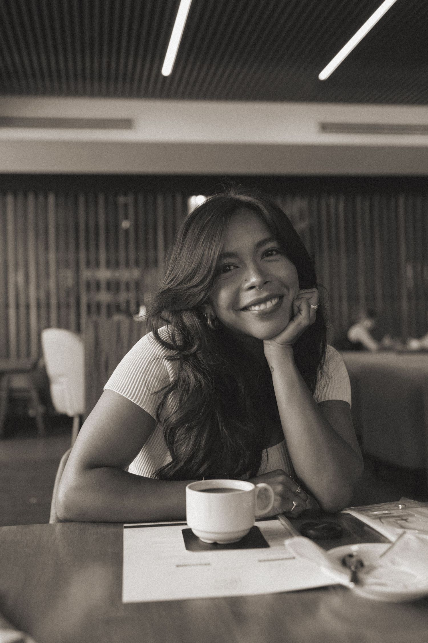 Smiling young woman resting chin on hand at cafe table with coffee cup in black and white photo.