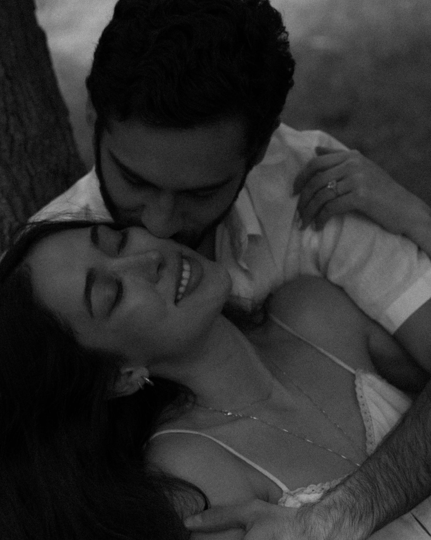 A black and white photo of a couple in an intimate embrace, the man kissing the smiling woman near a tree.