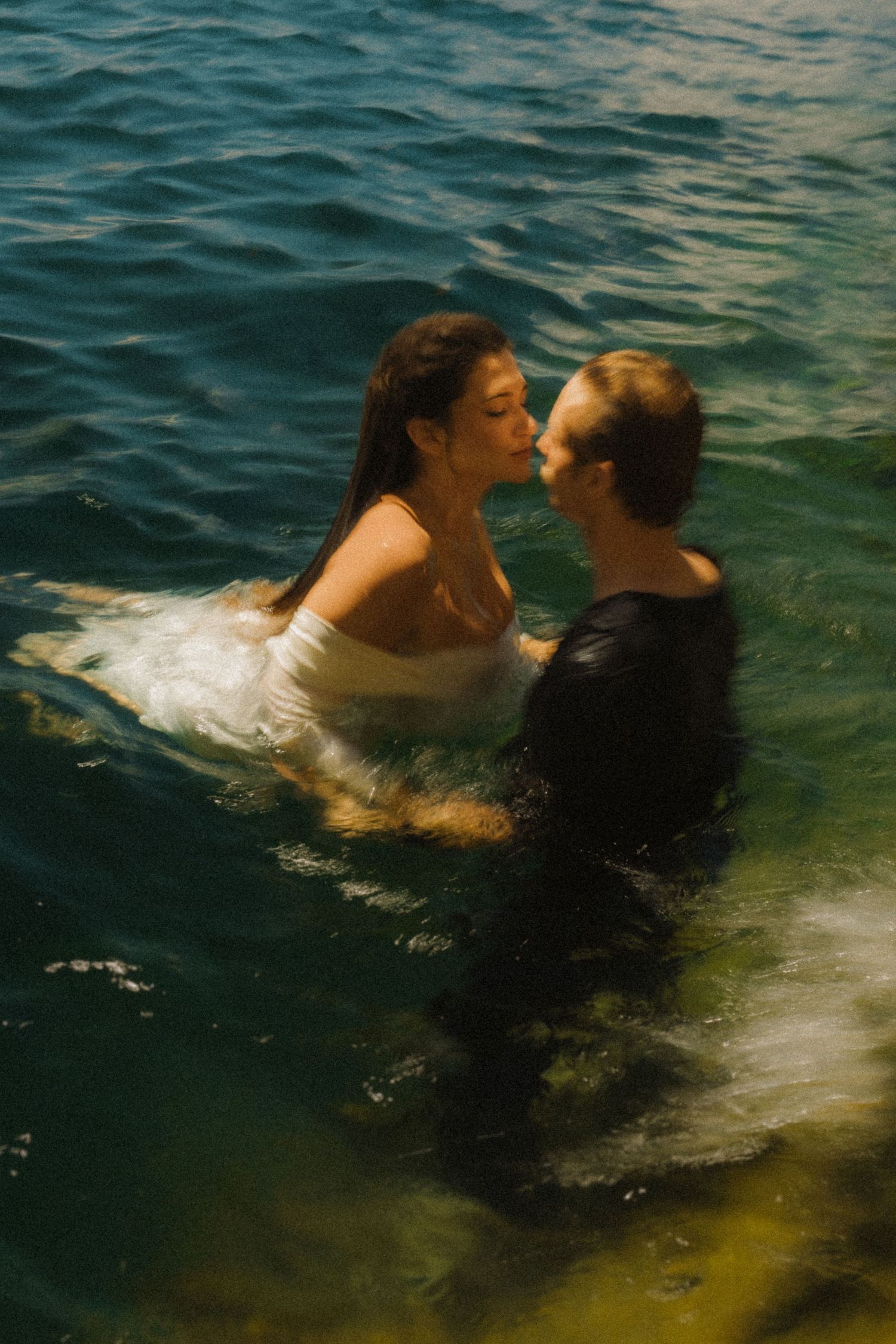 Romantic couple sharing a kiss while standing in turquoise water, wearing white and black outfits.