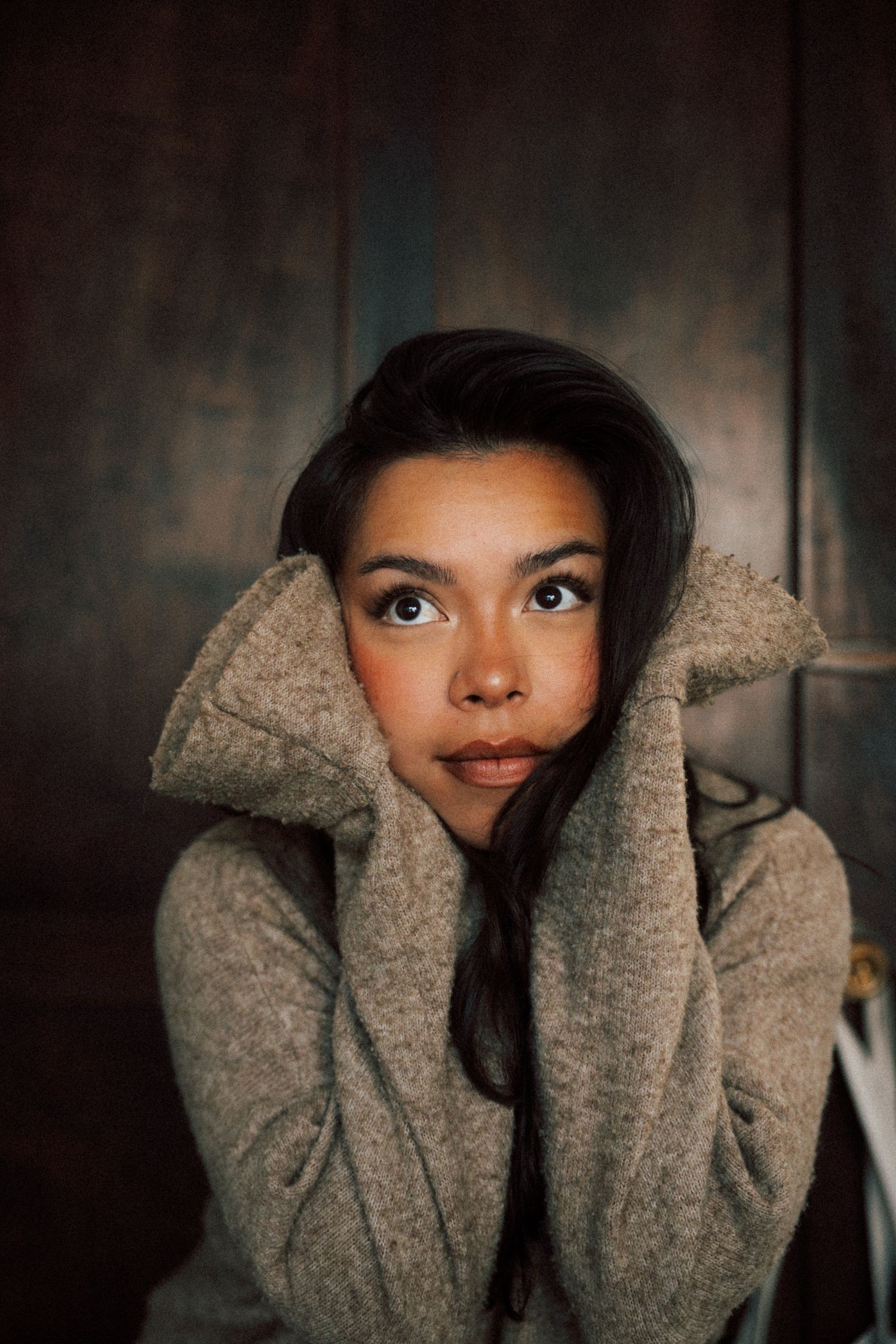Young woman in cozy gray wool sweater hugging collar close to face with expressive eyes against dark wood background.