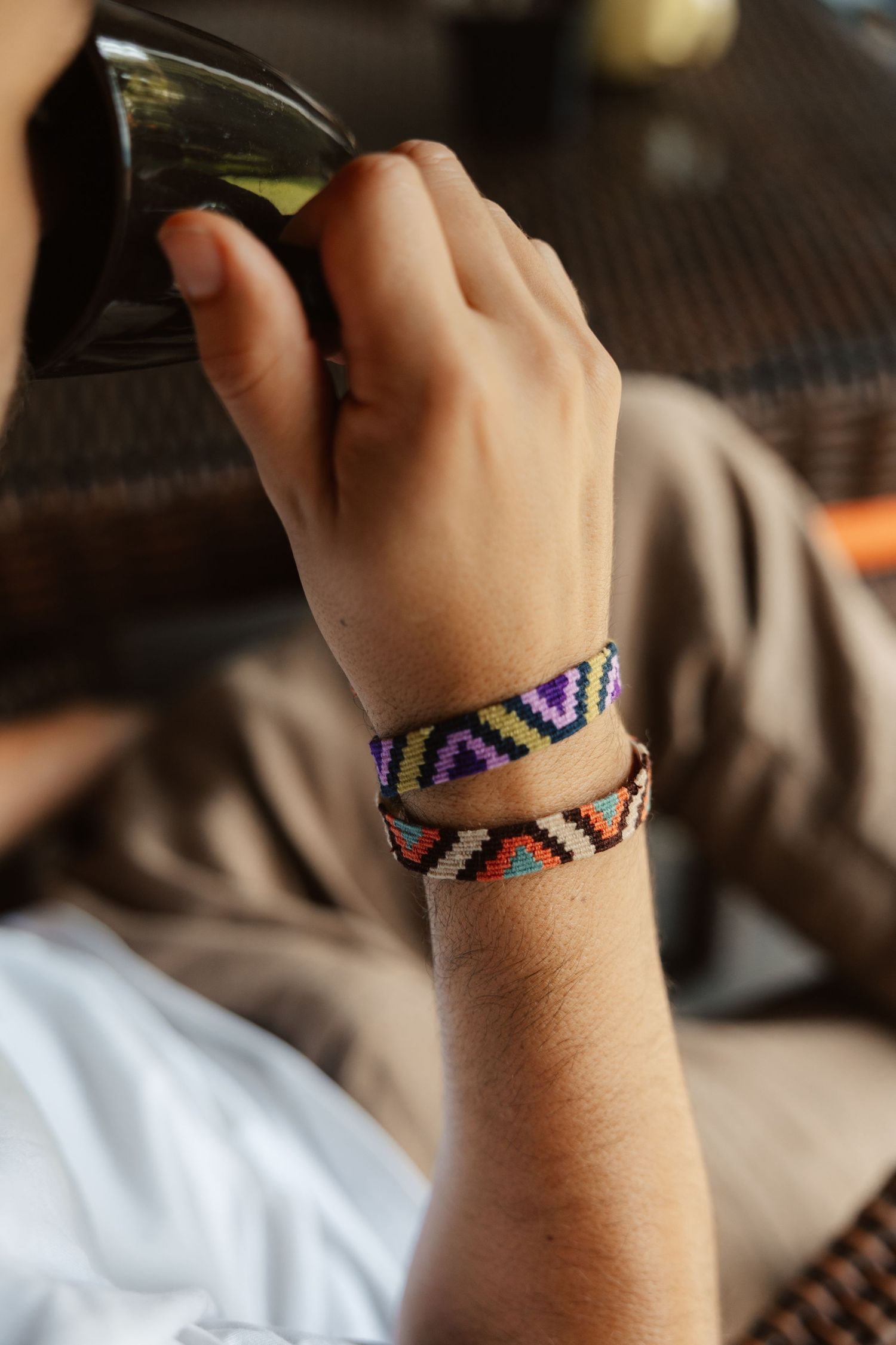 Close-up of wrist wearing colorful woven friendship bracelets with geometric patterns while holding a glass drink.