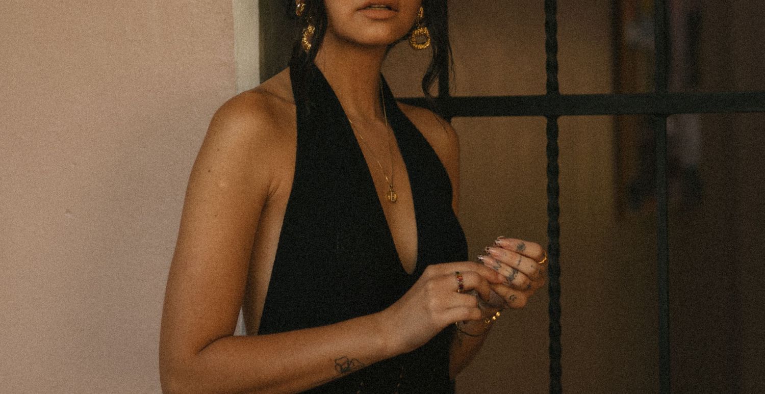 Woman in a black halter dress with gold jewelry standing near a window in natural light.