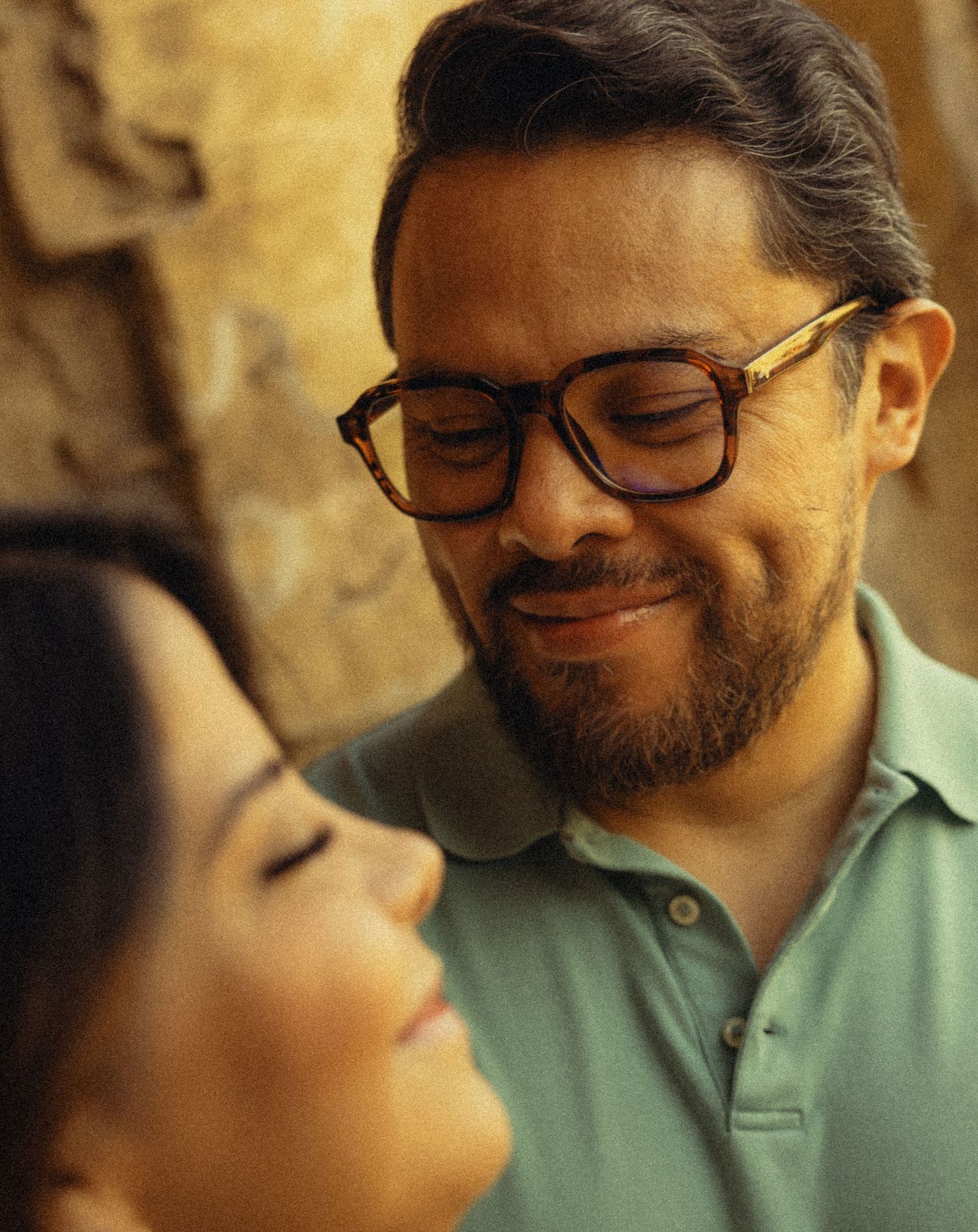 A bearded man with glasses smiles warmly at a woman with closed eyes in a tender, intimate moment.