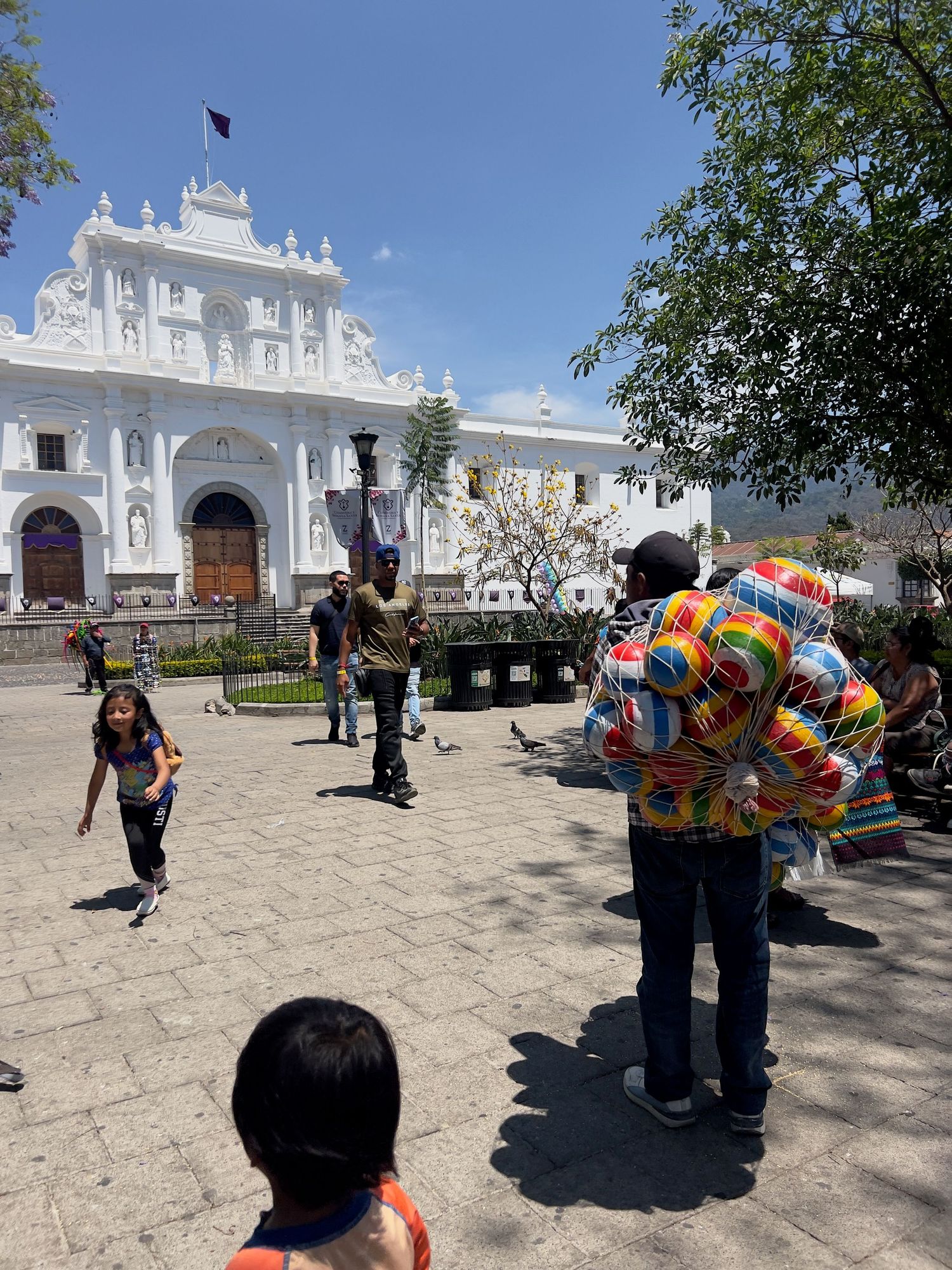 Balloon vendor selling colorful balloons in a sunny plaza near a white colonial church in Antigua, Guatemala.
