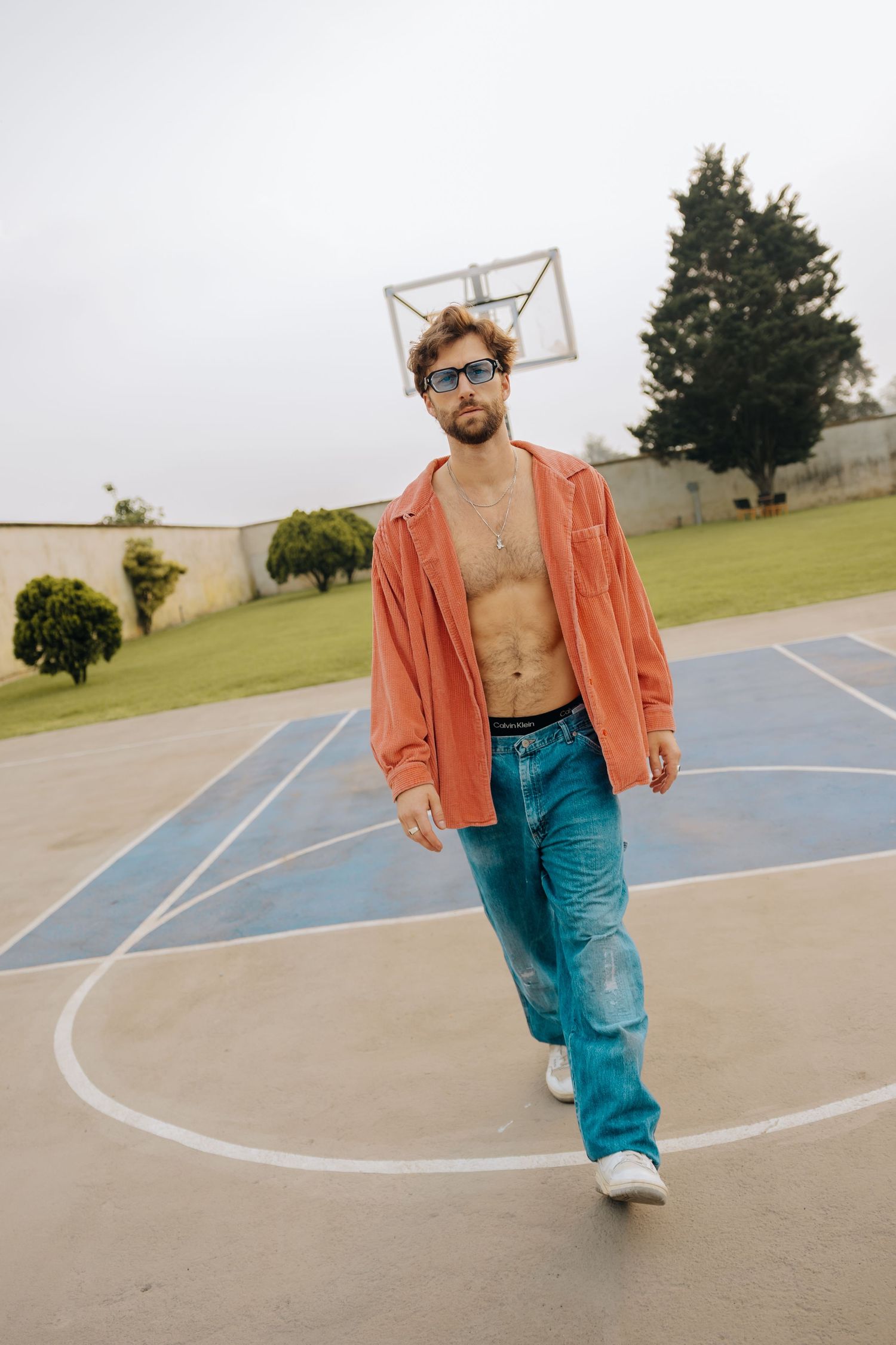 Stylish man in open orange jacket, blue jeans, and sunglasses walking on an outdoor basketball court.