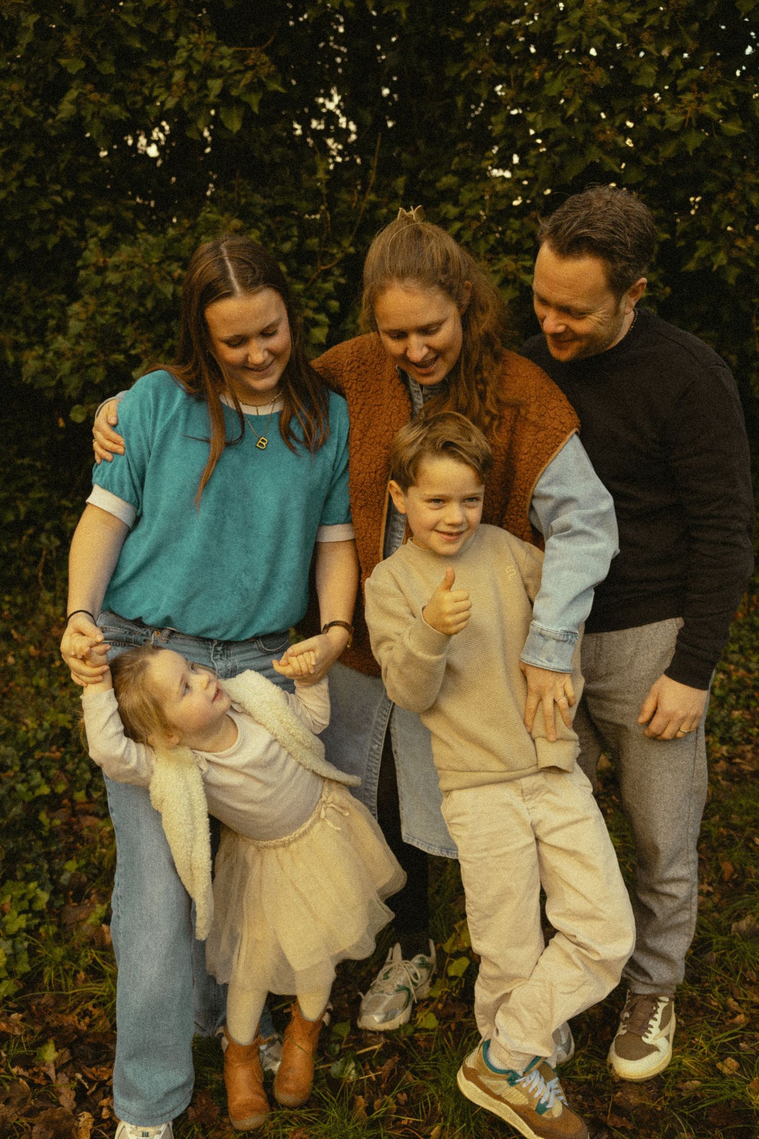 A happy family of five poses together outdoors, with a boy giving thumbs up among green trees.