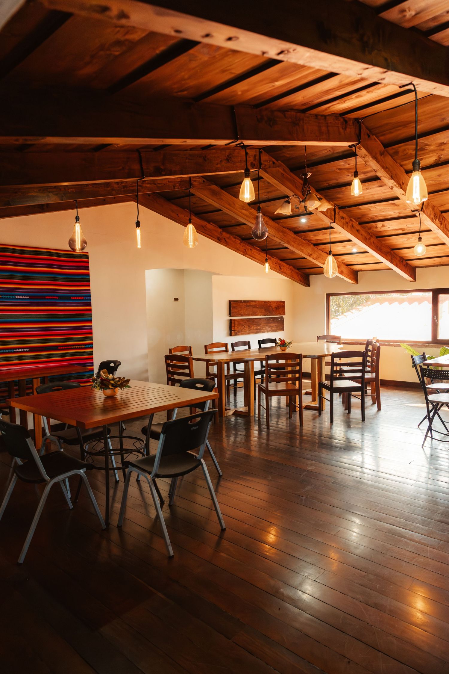 Rustic restaurant interior with wooden ceiling beams, Edison bulb pendant lights, dark hardwood floors, and colorful textile wall art.