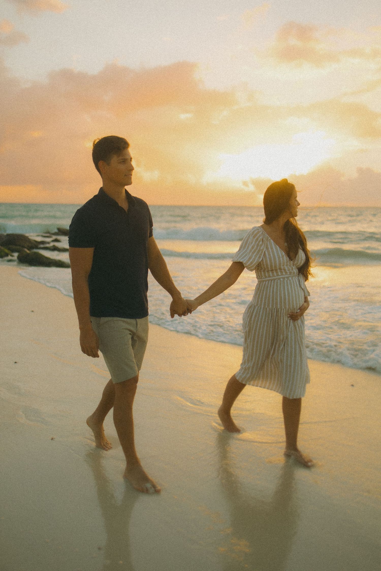 Pregnant couple holding hands walking barefoot on a beach at sunset, woman in striped dress touching baby bump.