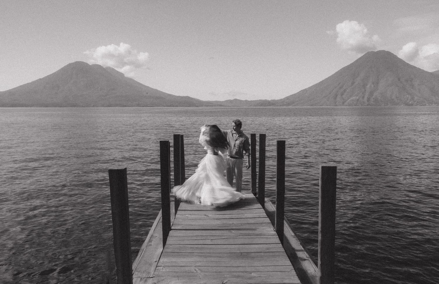 Black and white photo of bride and groom on wooden dock with volcanic mountains over lake.