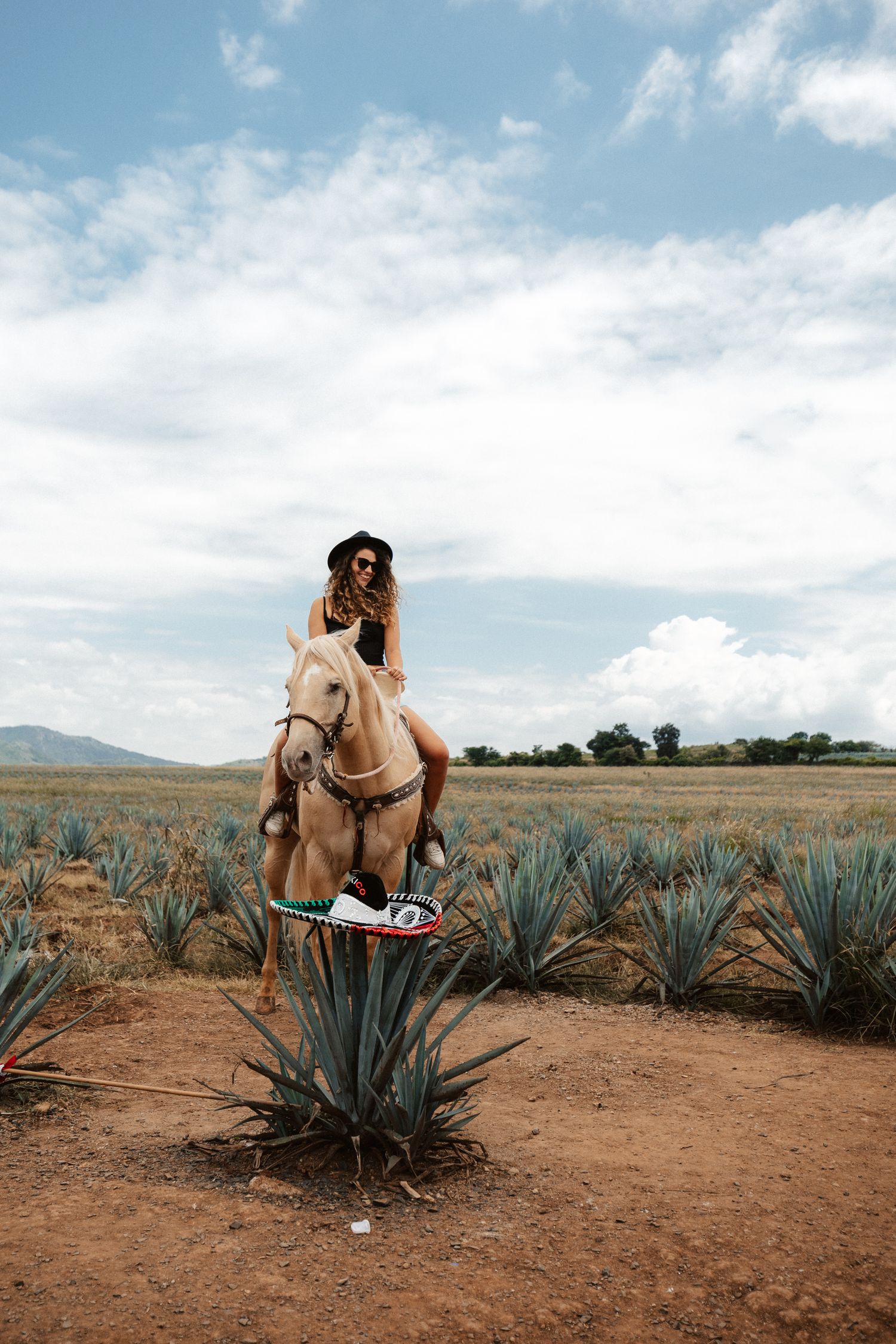Woman in black hat riding a palomino horse through an agave field under a cloudy sky in Mexico.