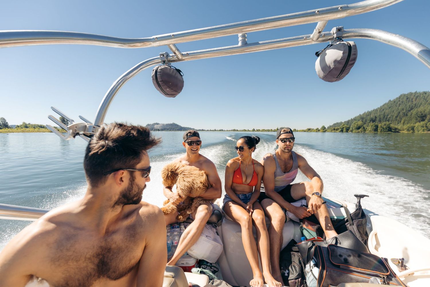 Four friends enjoying a sunny boat ride on a calm river surrounded by lush green trees on a clear day.