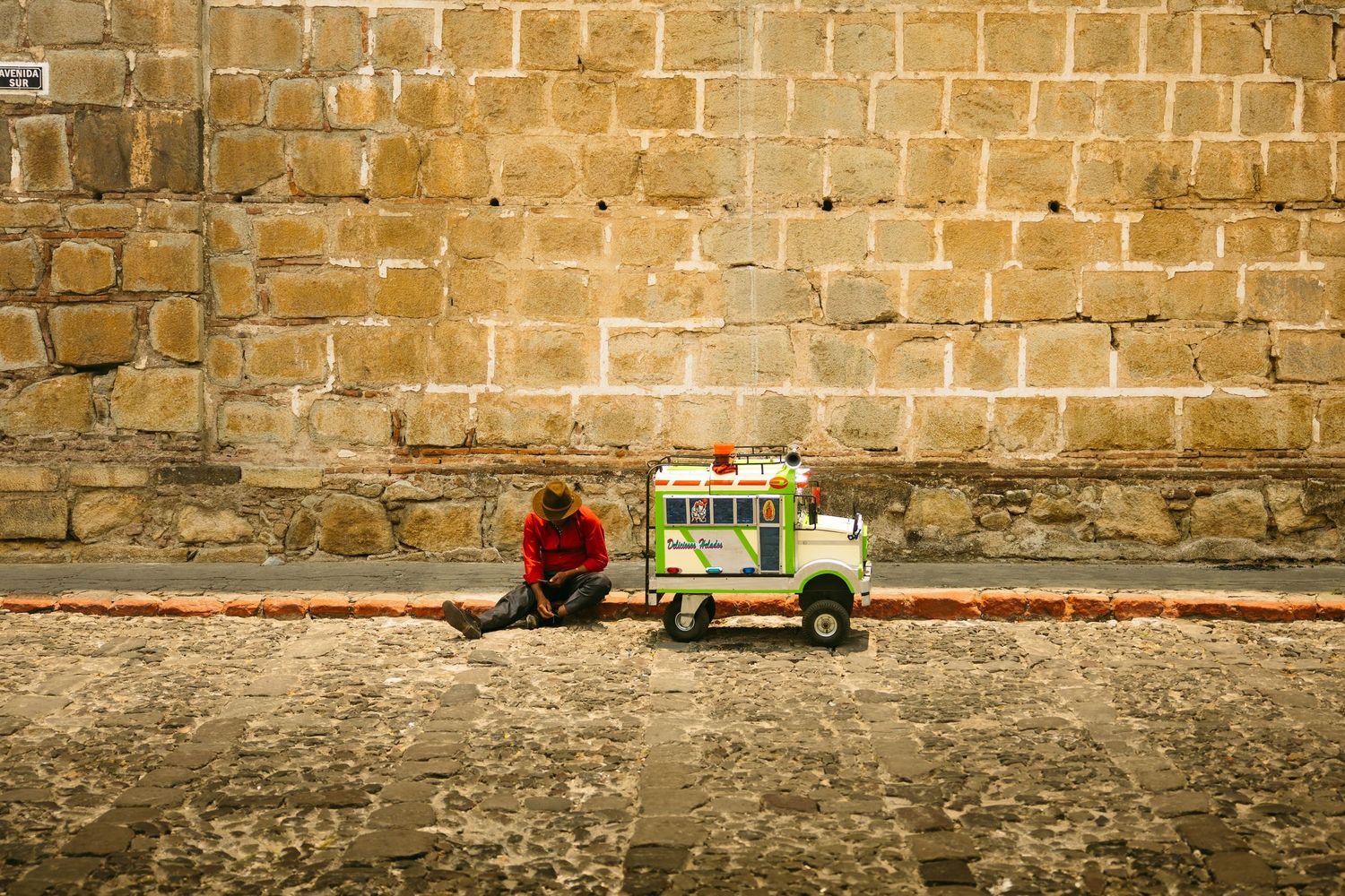 Person in red sitting beside a colorful food cart against a large stone wall on a cobblestone street.