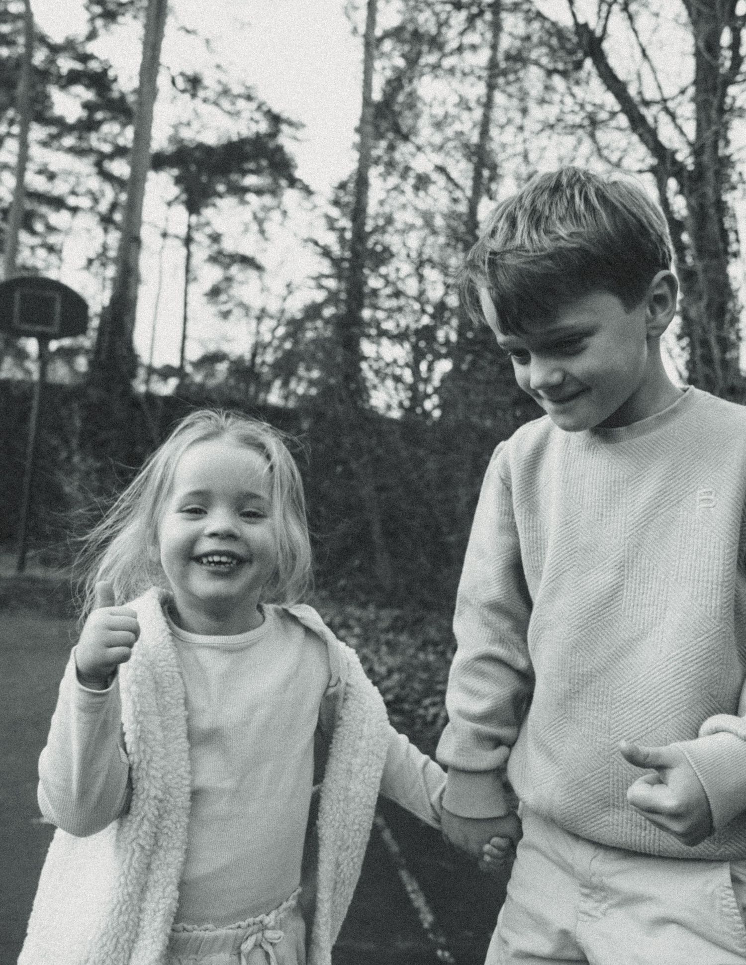 Two smiling children holding hands outdoors in a black and white photo, with trees and a basketball hoop in background.