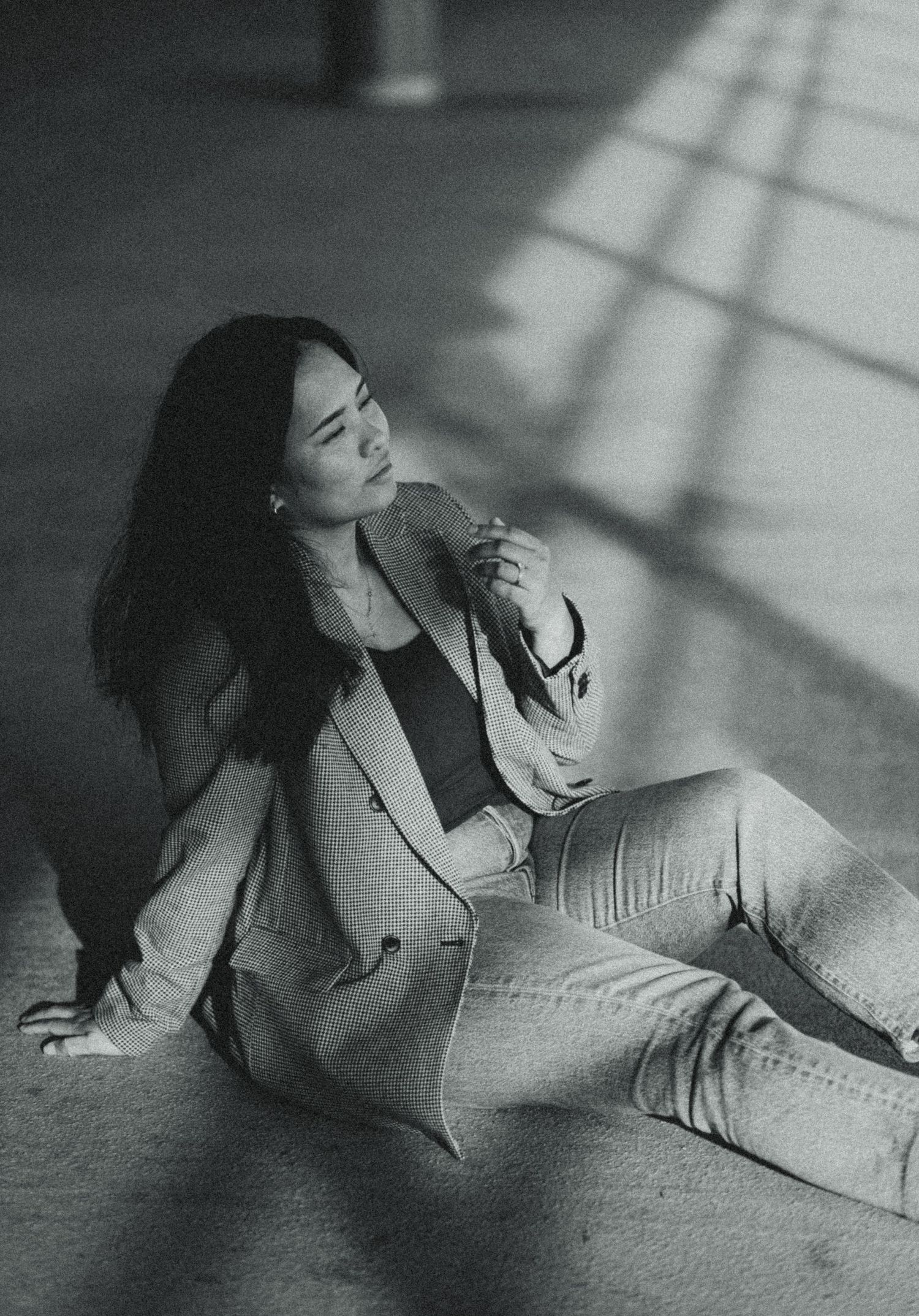 Black and white photo of a young woman in blazer and jeans sitting on floor with dramatic shadow patterns.