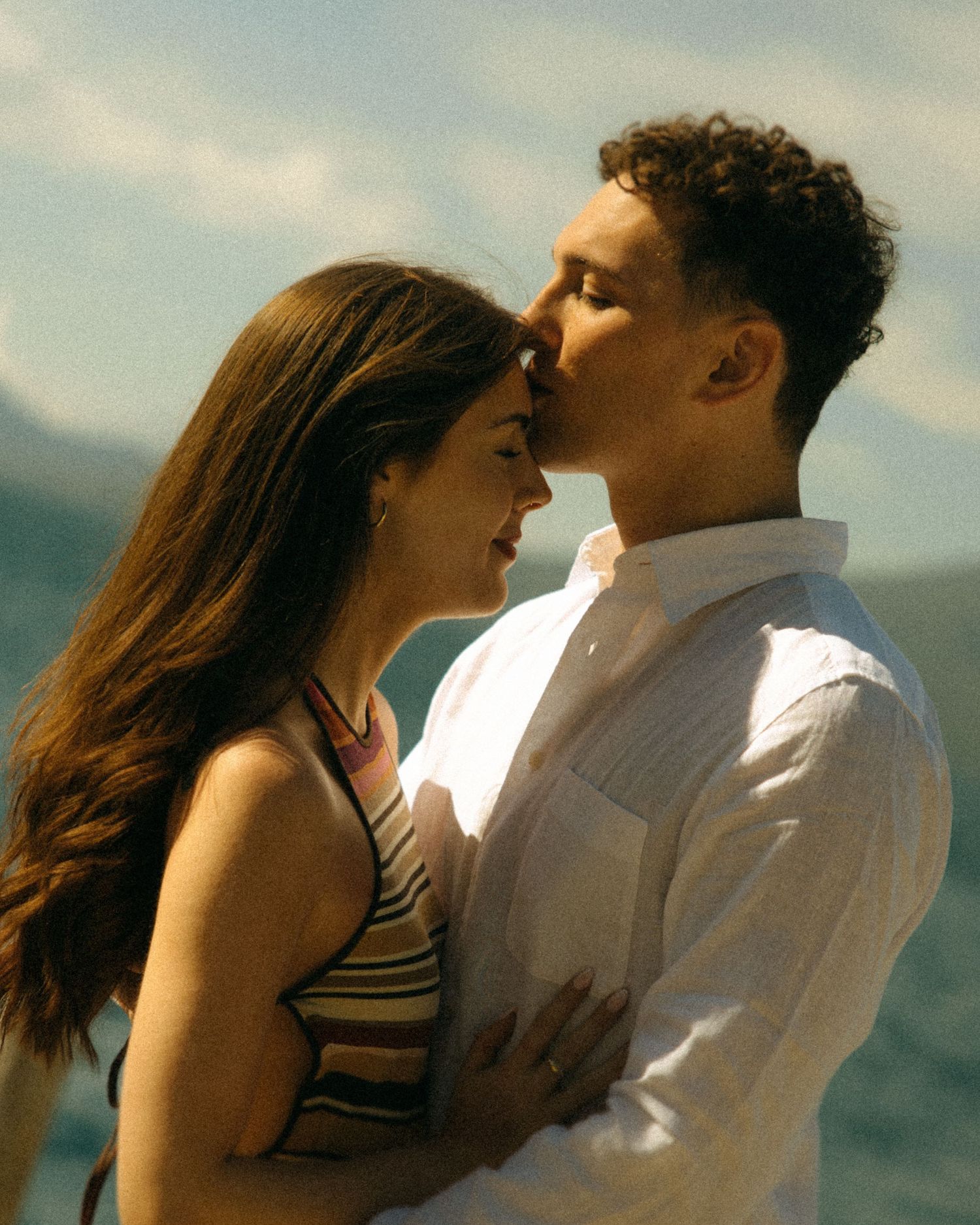 A romantic couple shares an intimate moment by the water, with mountains in the background on a sunny day.