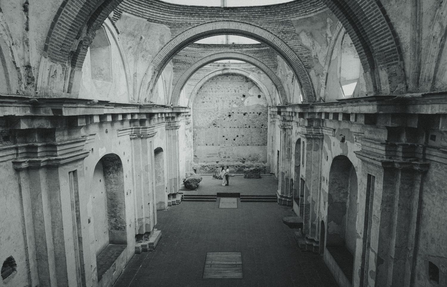 Black and white interior of a ruined colonial church with tall stone arches, weathered walls, and an open roof leading to a distant altar.