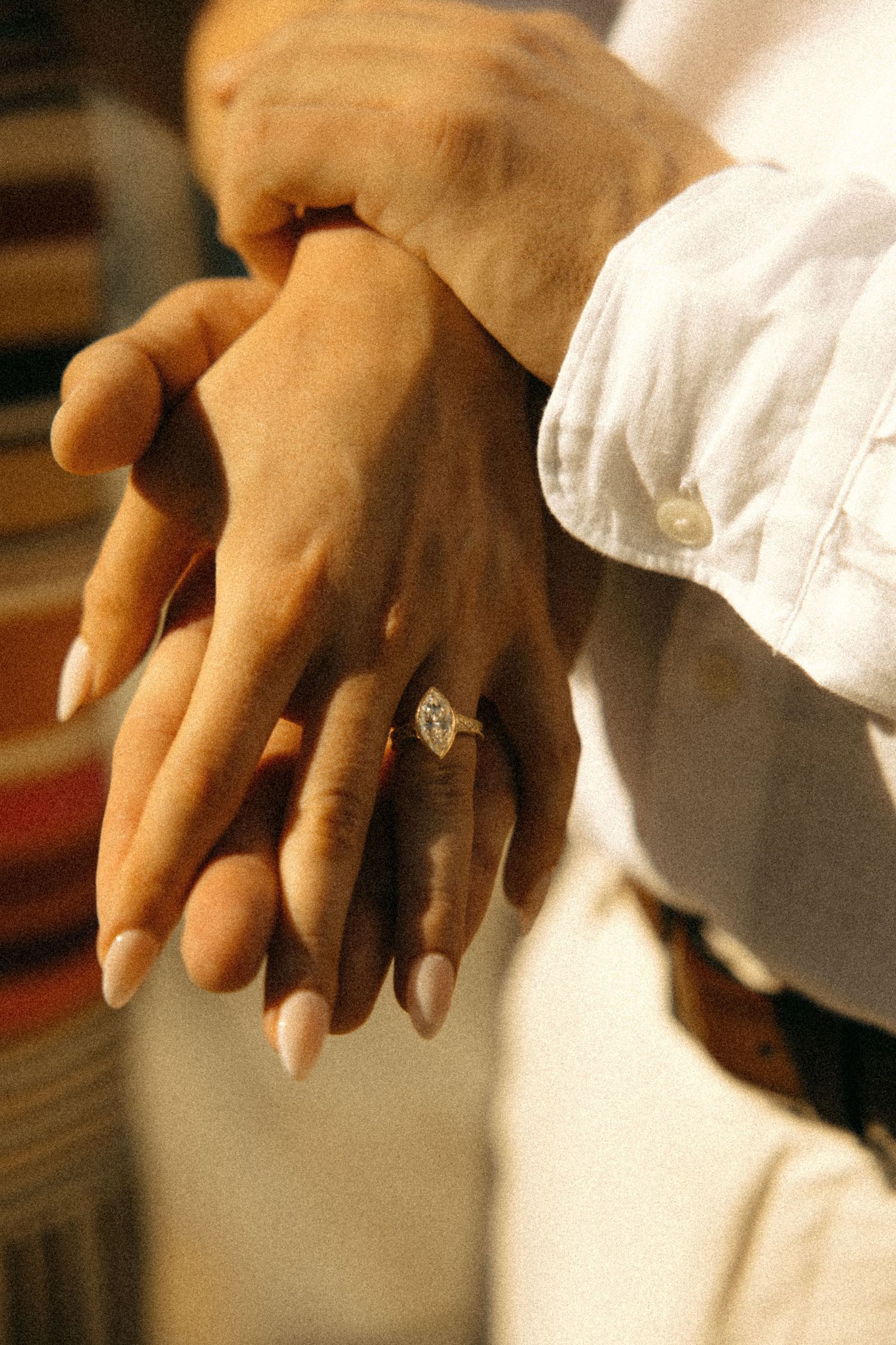 Close-up of two hands intertwined, one wearing a marquise diamond engagement ring, with white shirt sleeve visible.