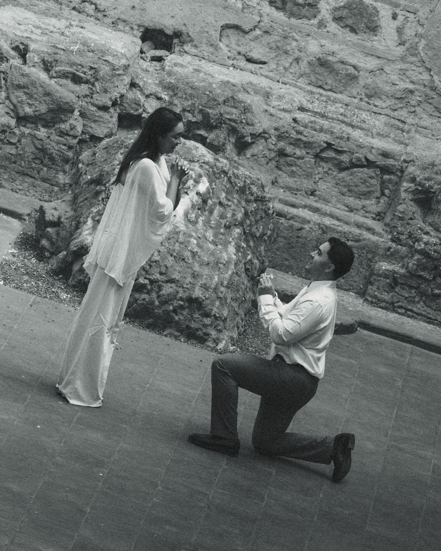 Black and white photo of a man on one knee proposing to a woman in a white dress near a stone wall.