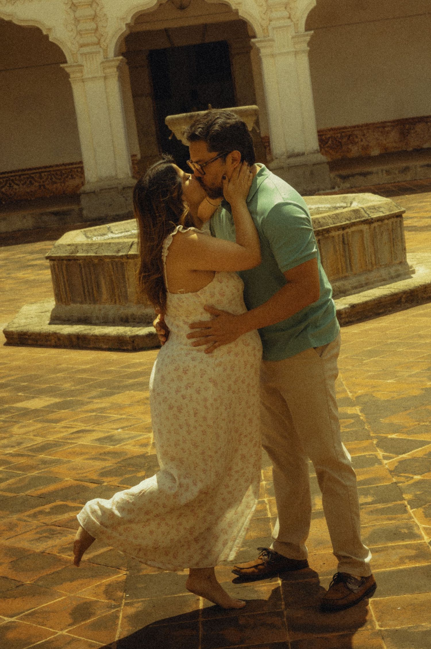 A couple sharing a romantic kiss in a sunlit courtyard with arched architecture.
