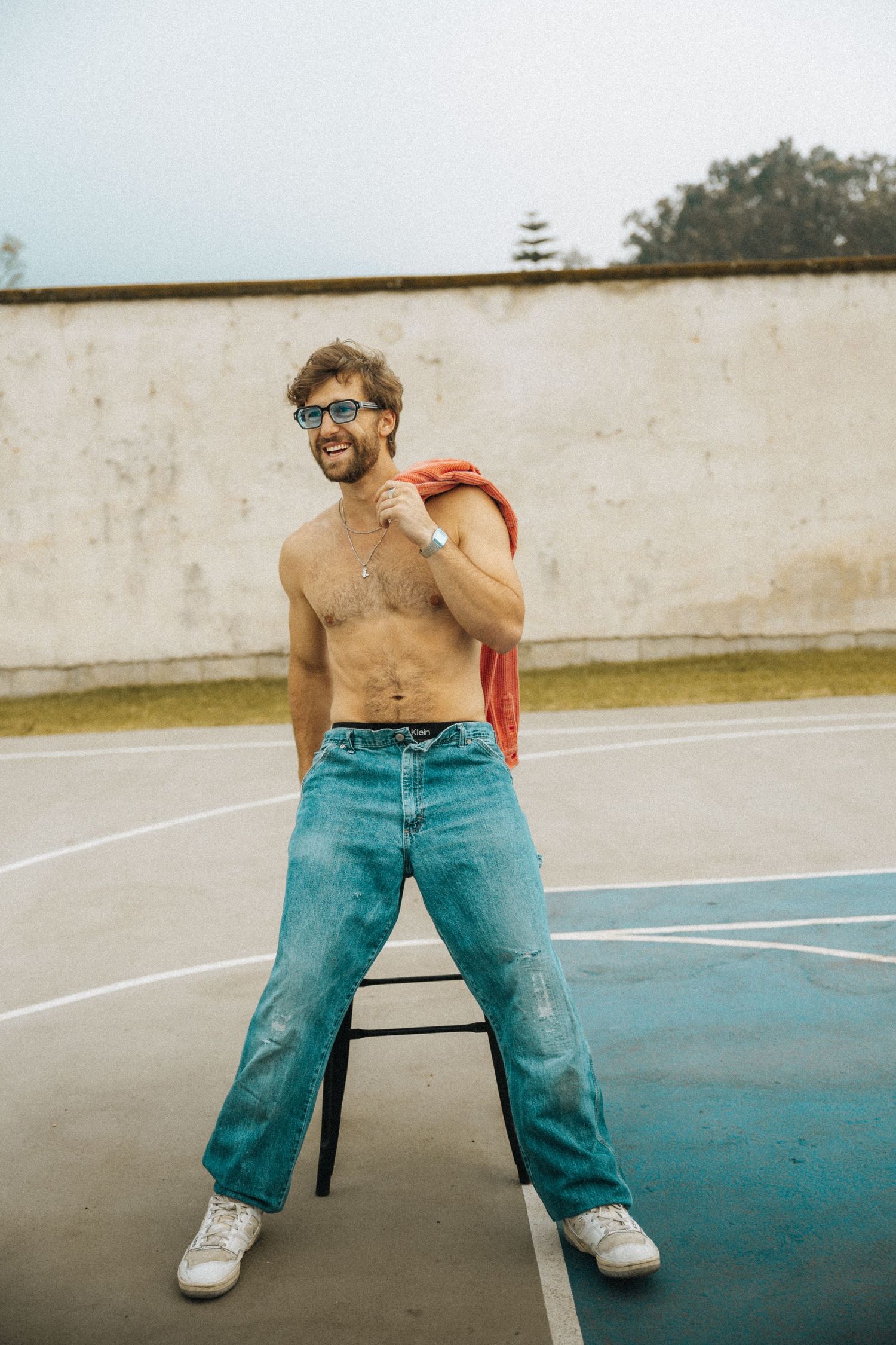 Shirtless man in blue jeans and sunglasses posing on a chair at an outdoor basketball court.