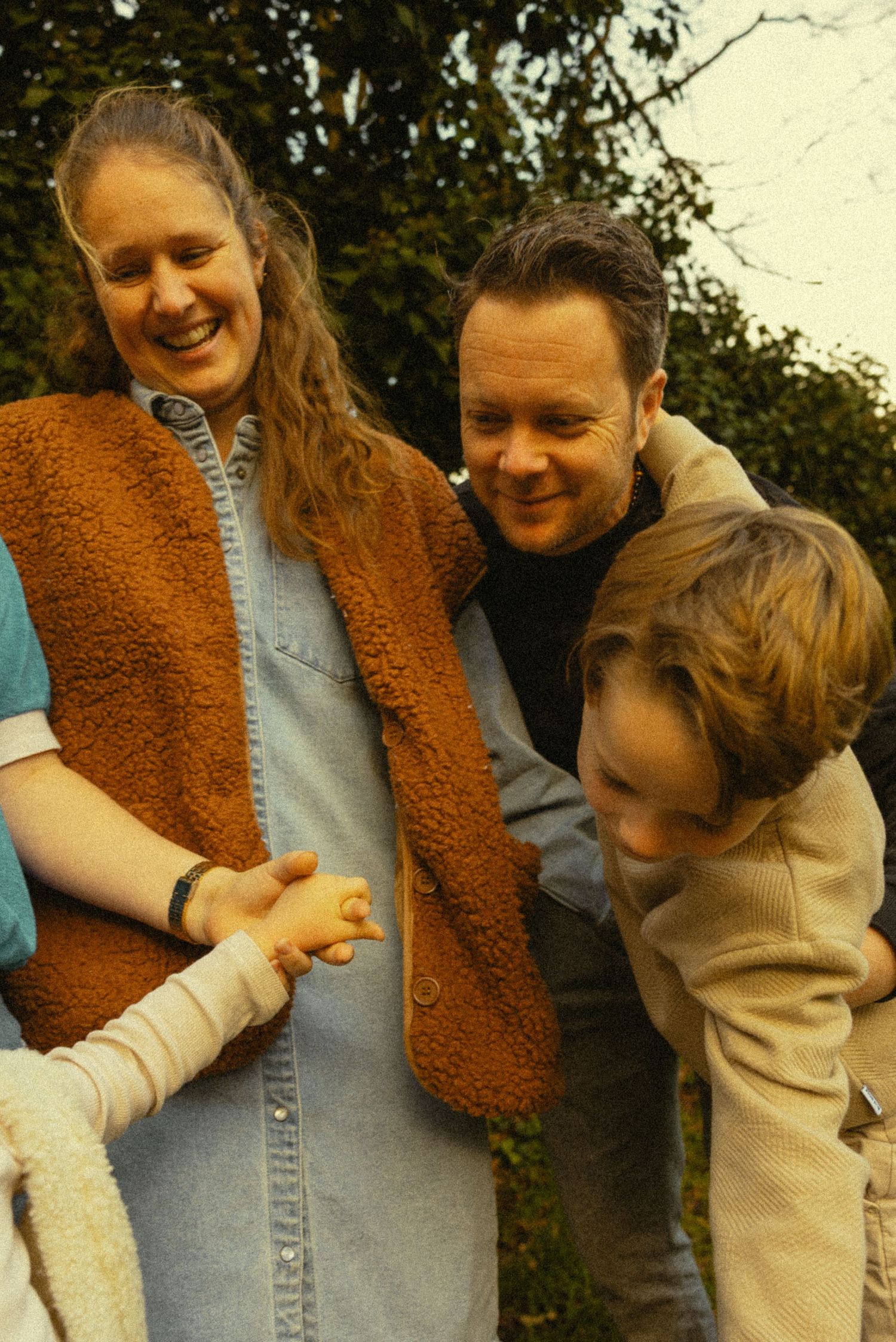 A smiling family with a woman in a brown vest, a man, and a child gathered together outdoors near green trees.