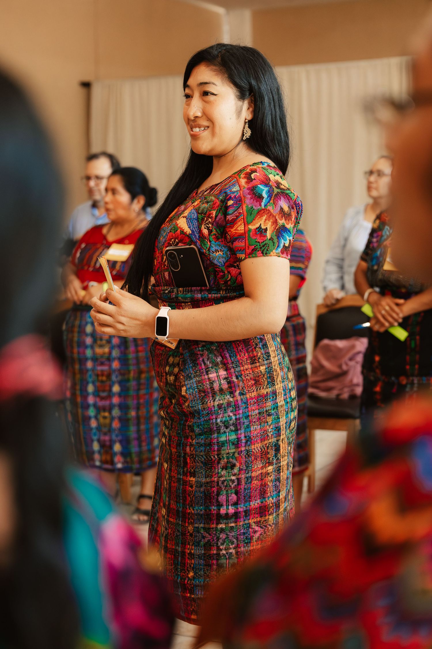 Woman in vibrant traditional Guatemalan huipil dress smiling at indoor cultural gathering.