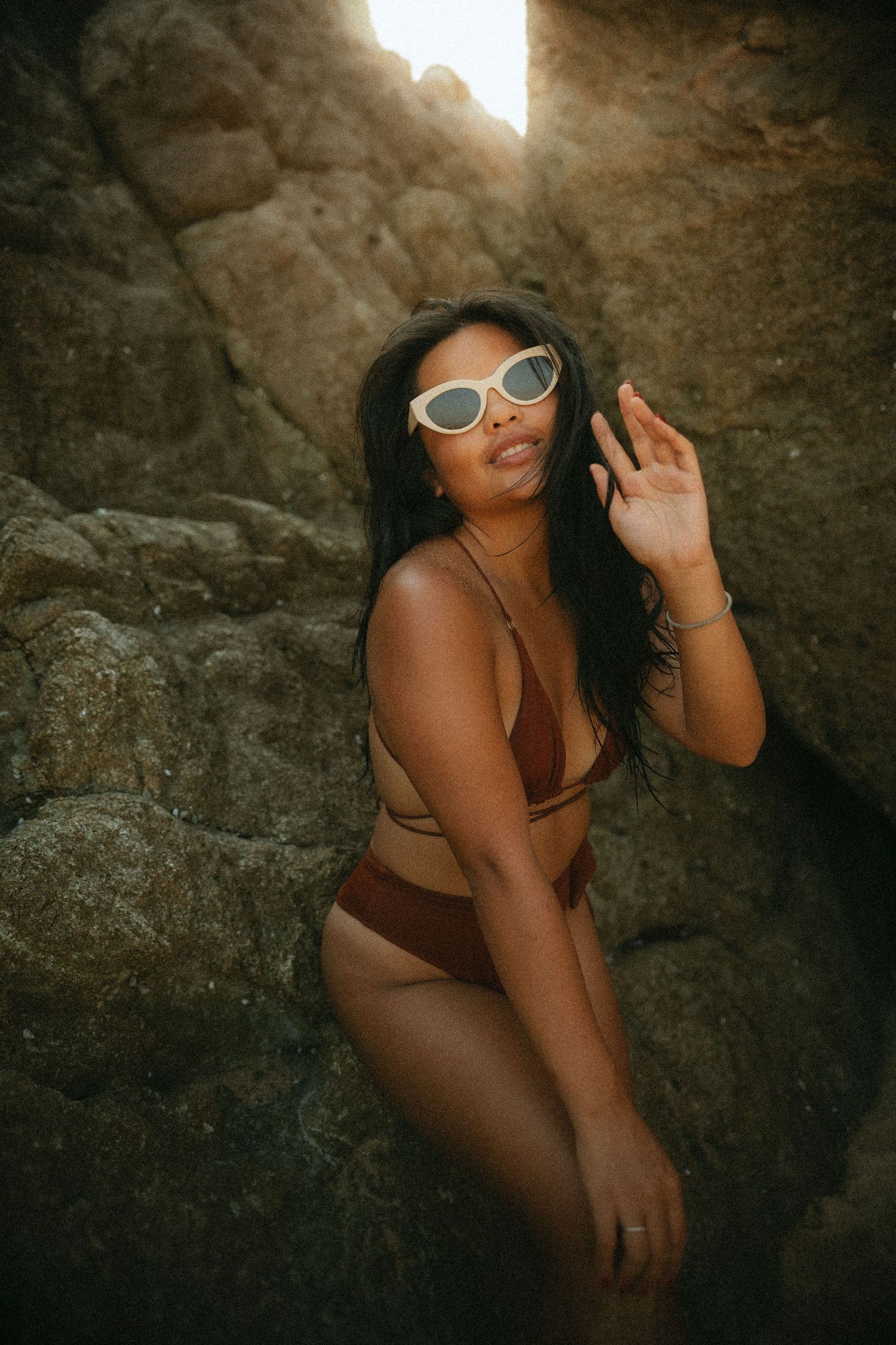 Woman in brown bikini and white sunglasses posing between rocky formations with sunlight streaming through.
