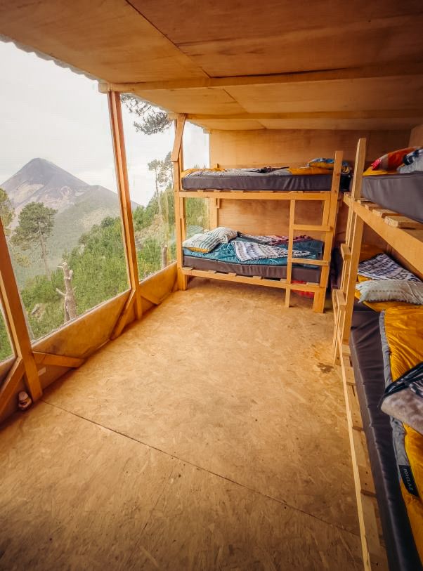 Rustic wooden cabin interior with bunk beds and panoramic mountain views through large glass windows.