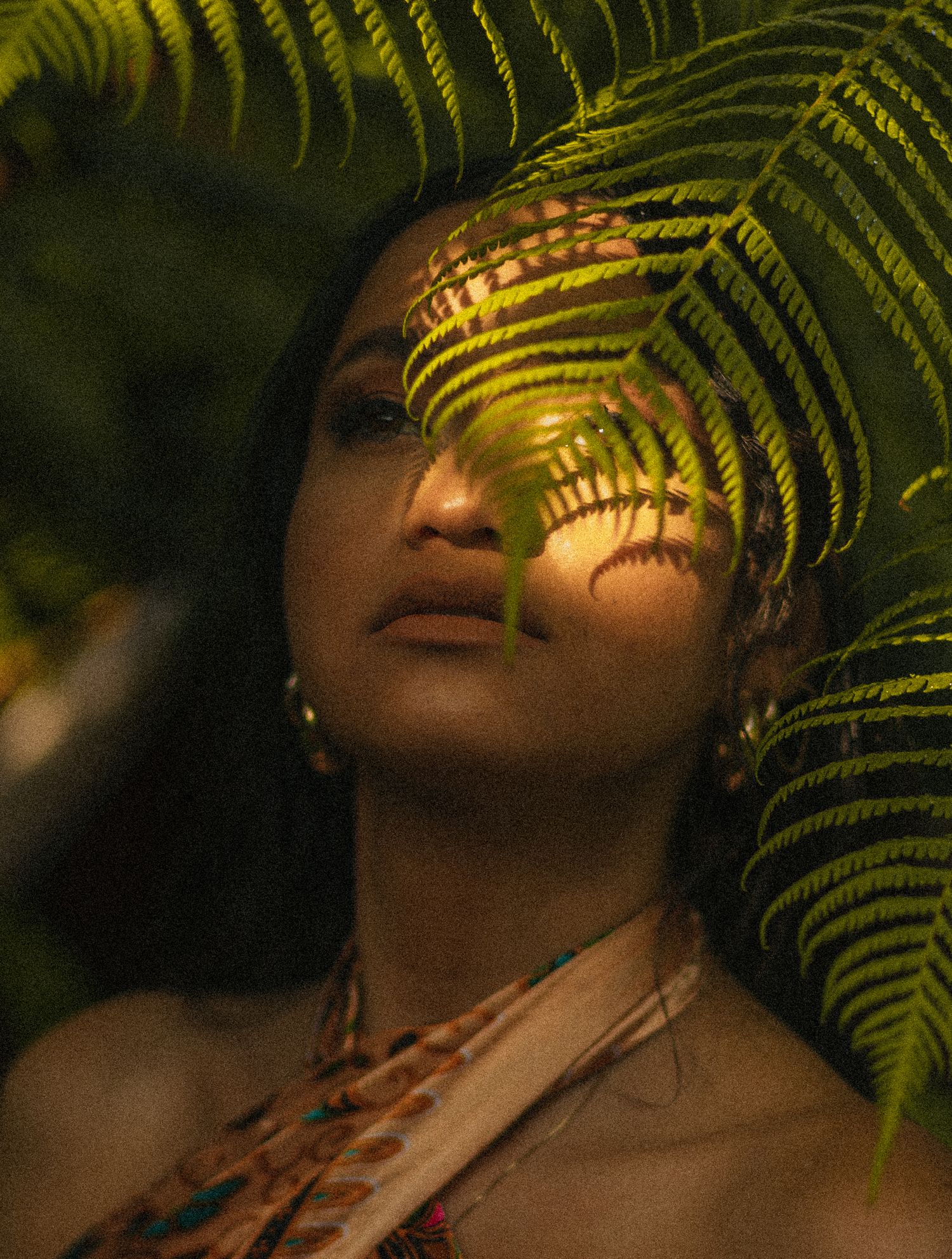Young woman gazing upward through lush tropical fern leaves in a moody jungle setting.