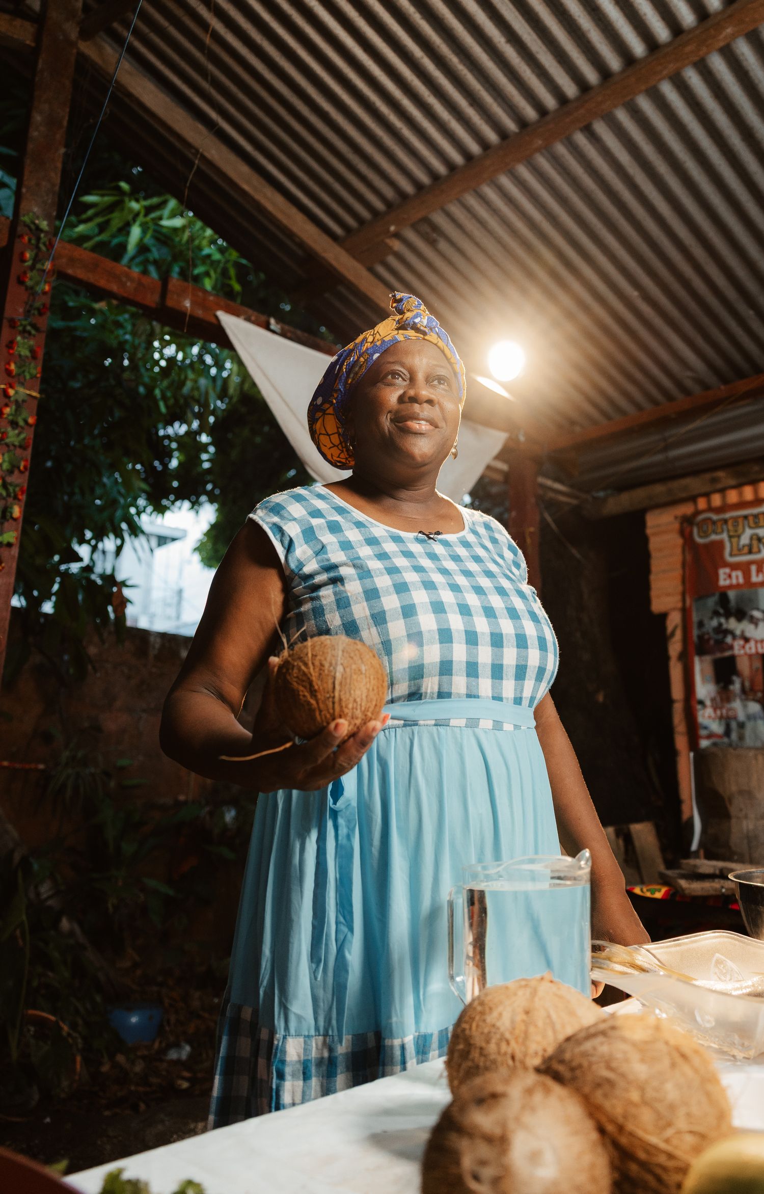 Smiling African woman in blue gingham dress holding coconut at outdoor market stall.