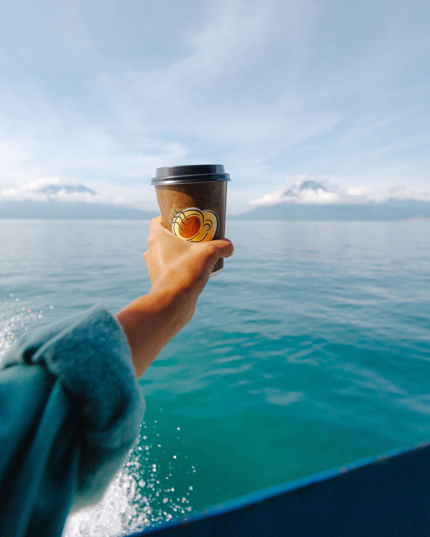 Hand holding a dark coffee cup with golden logo on a boat with turquoise ocean and snowy mountains backdrop.