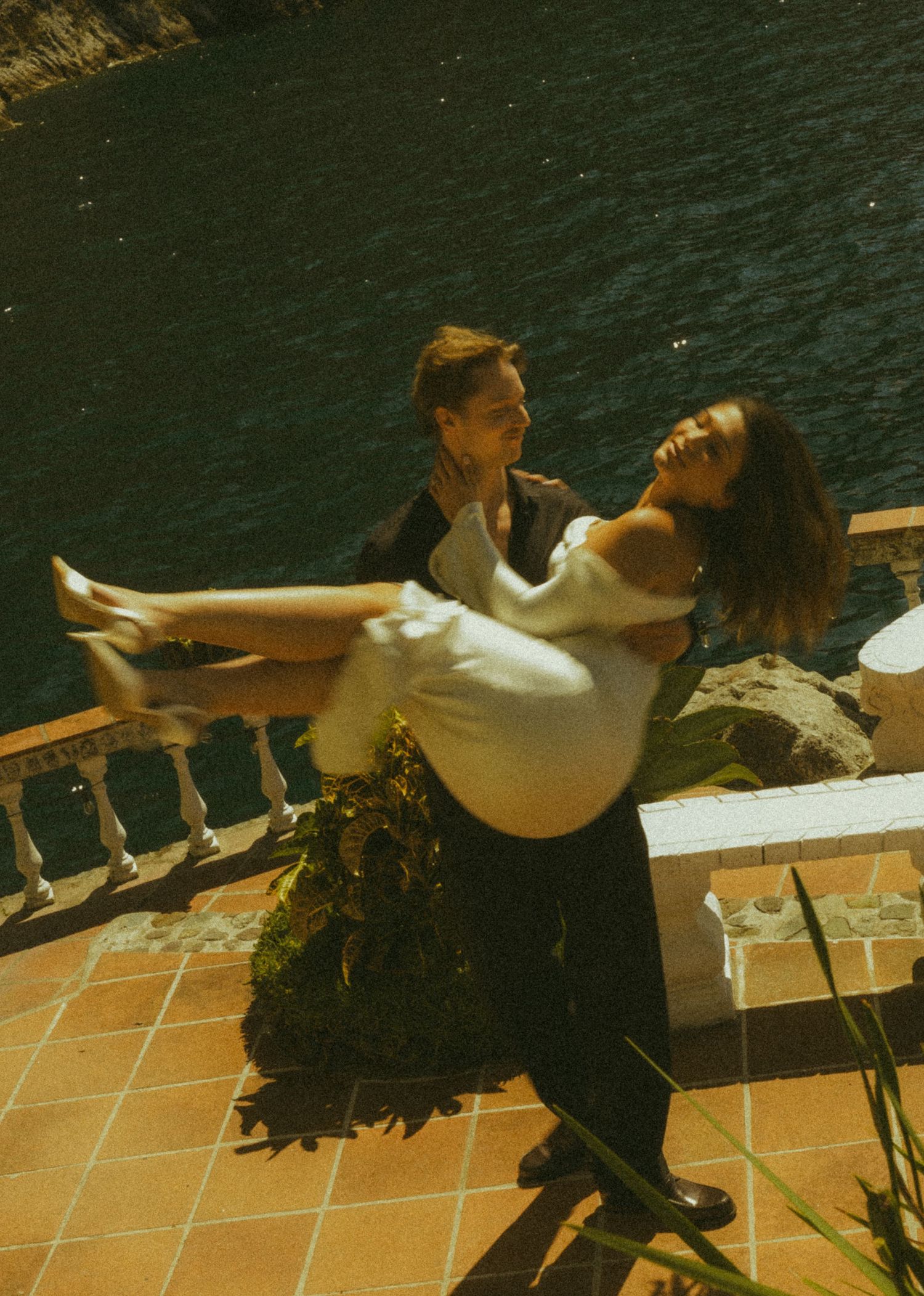 Groom carrying laughing bride in white dress on sunlit terrace overlooking turquoise water.