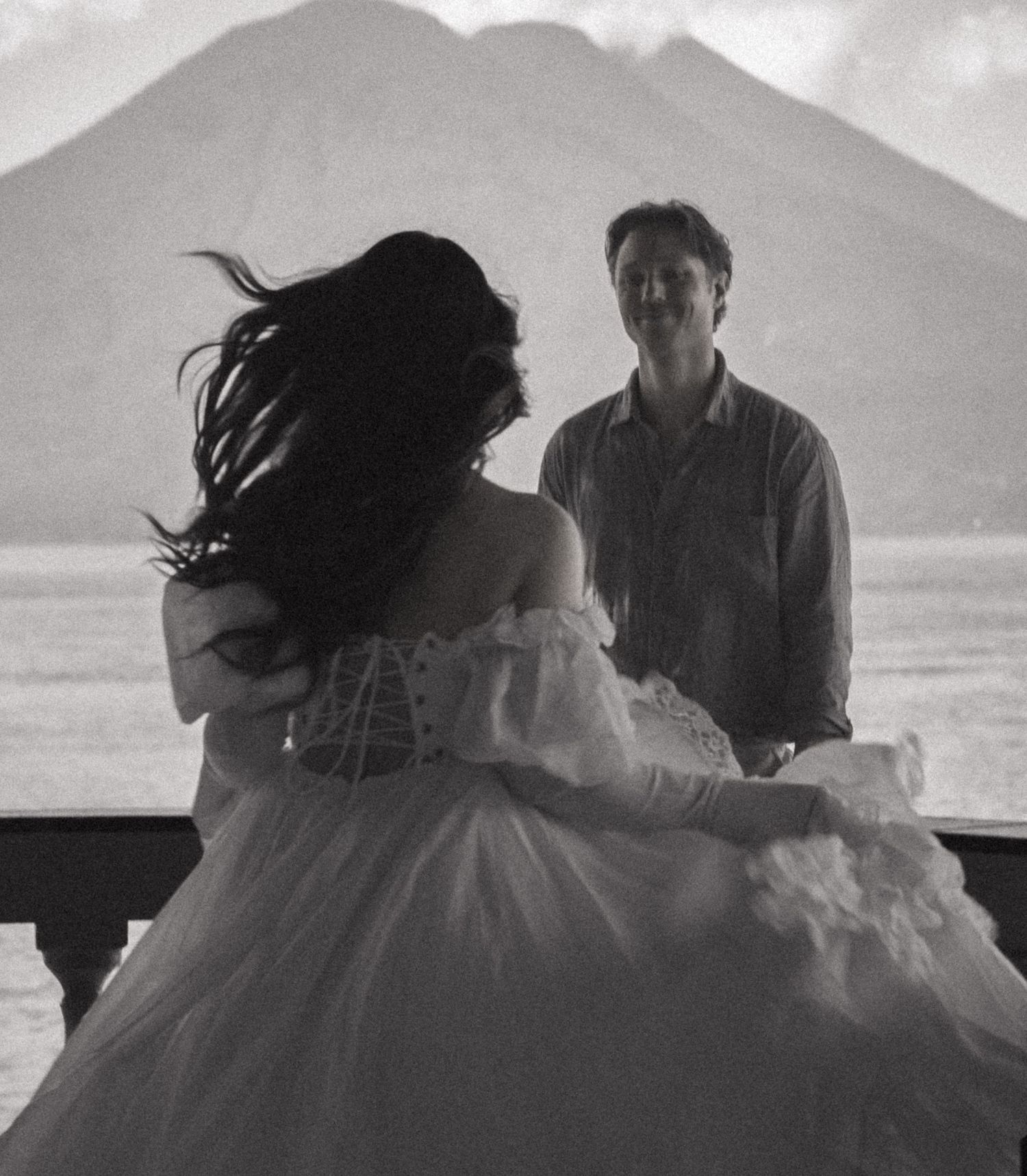 Black and white photo of a bride in a ball gown twirling before a groom with volcanic mountains behind.