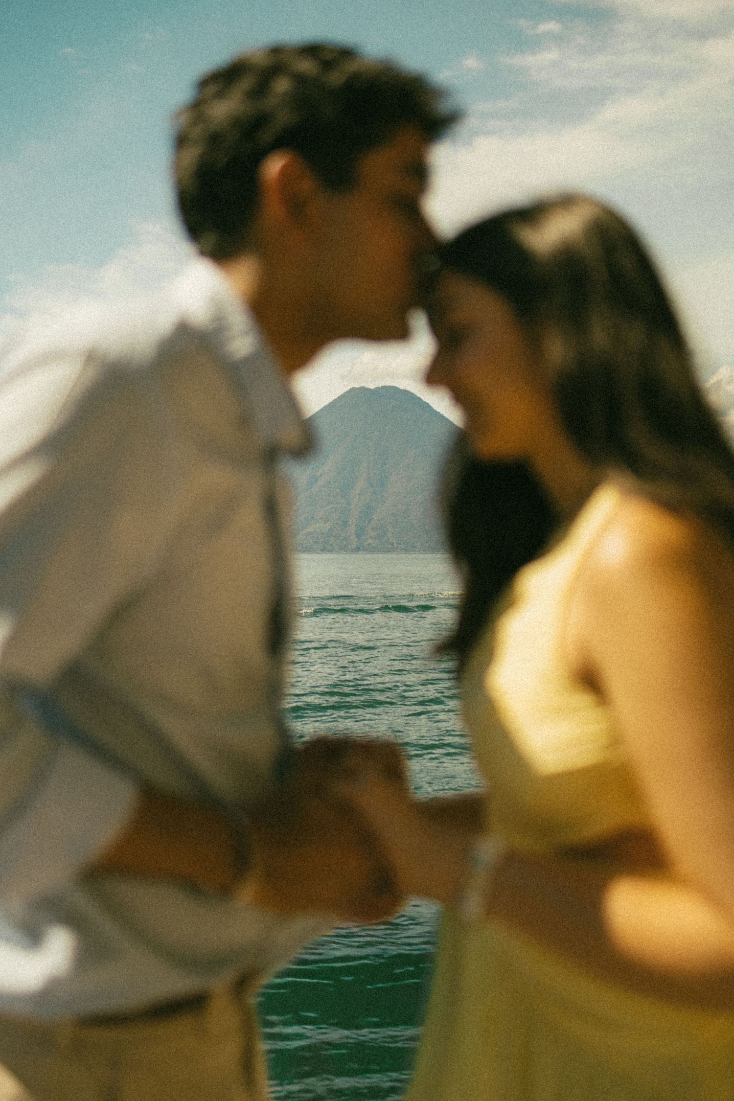 A blurred romantic couple holding hands as he kisses her forehead beside a sparkling lake with a mountain in the distance.