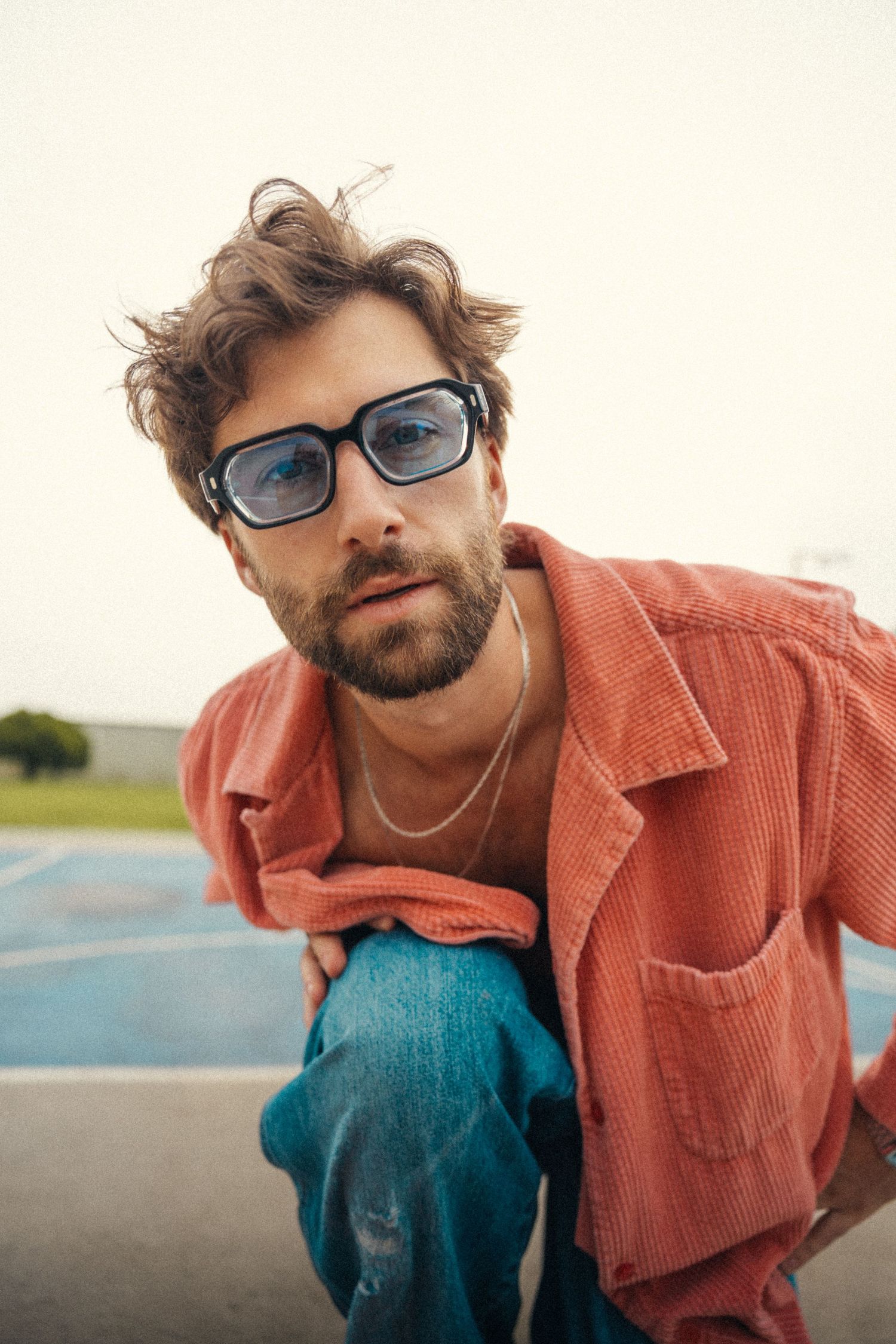 Bearded man with wavy hair wearing black glasses, coral corduroy shirt, and jeans, sitting outdoors on a blue surface.