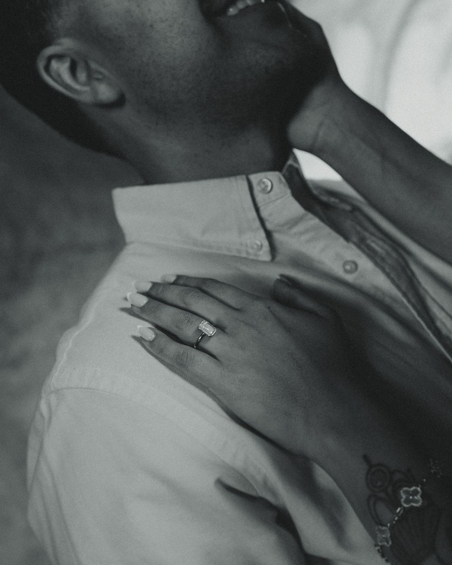 Black and white close-up of a woman's hand with a diamond engagement ring resting on a man's shoulder as he tilts his head back.