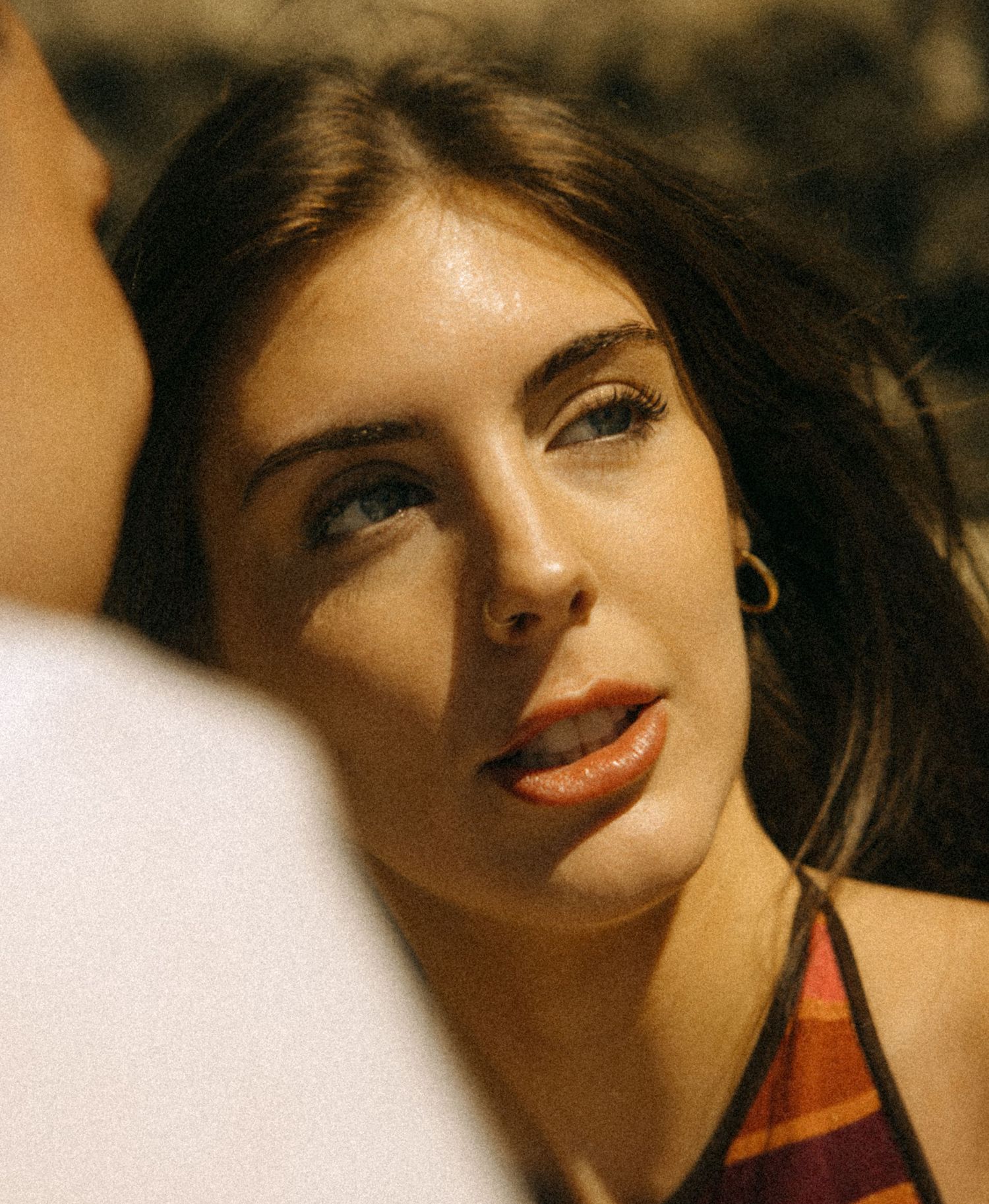 A young woman with dark hair and blue eyes wearing a plaid top and gold hoop earring looks up in warm sunlight.